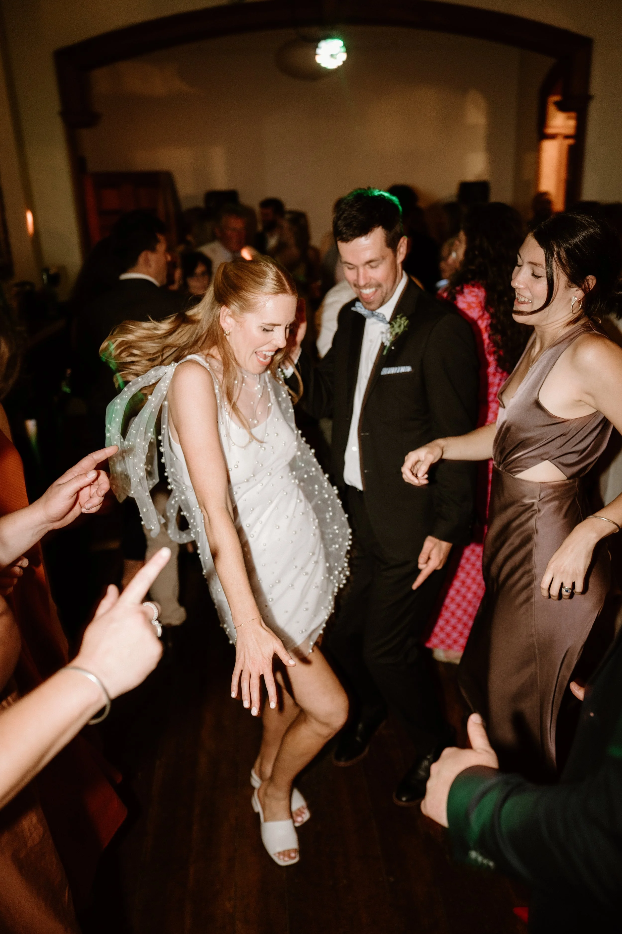 Group of people dancing at a celebration or wedding reception, with a woman in a white dress and a man in a tuxedo. Everyone appears to be happy and enjoying themselves.