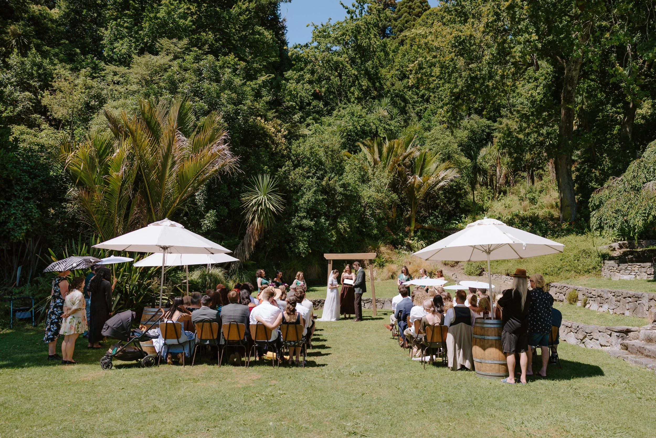An outdoor wedding ceremony with guests seated on chairs under white umbrellas, a couple exchanging vows, and a lush green forest in the background.