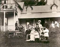 Black and white photograph of a group of people, including children and adults, posing in front of a house with a porch and a large yard.