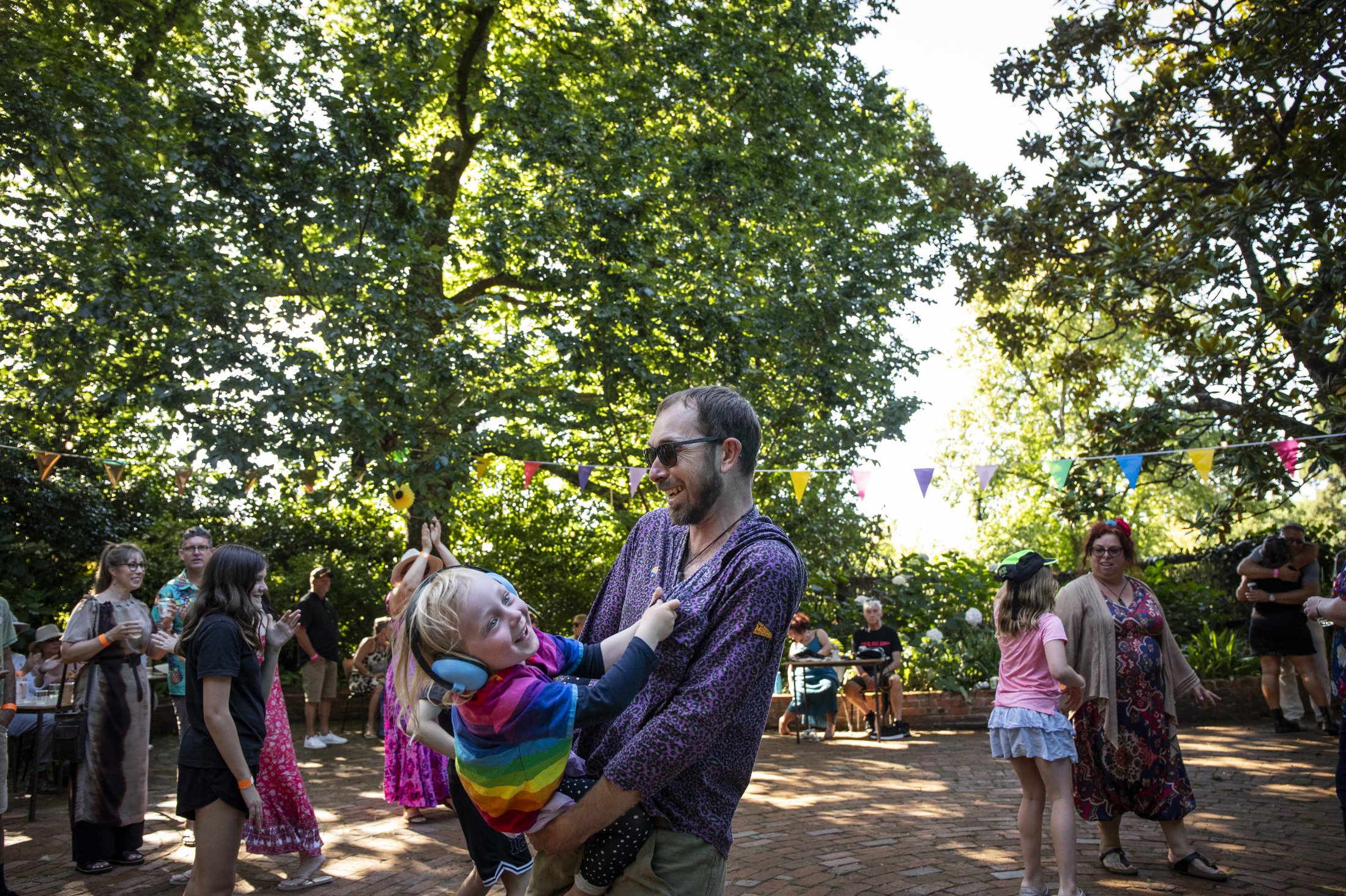 People gathered outdoors at a sunny festival with colorful bunting, trees, and cheerful interactions, including a man holding a young girl with rainbow-colored clothing.