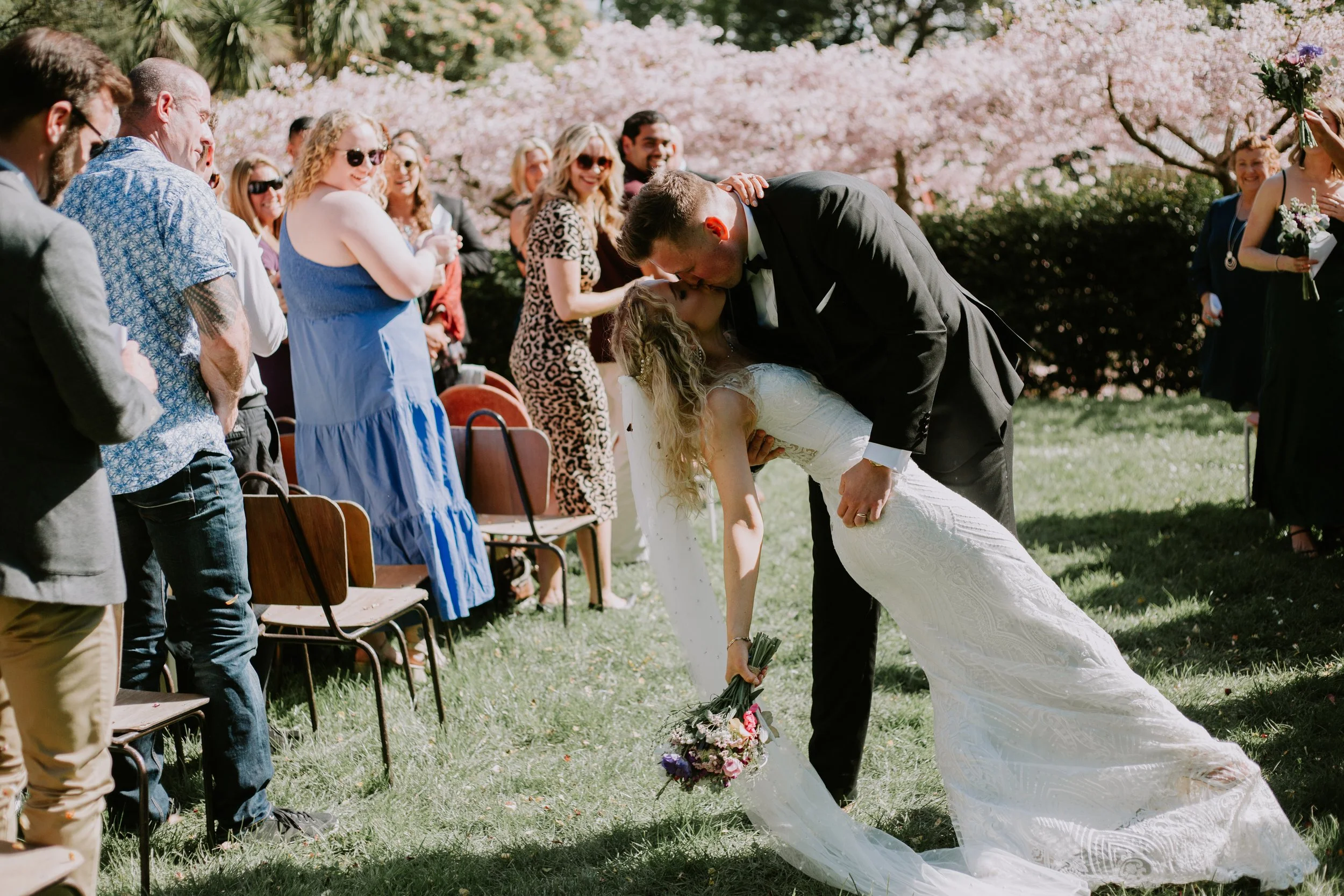 A bride and groom kiss during their outdoor wedding ceremony, surrounded by friends and family, under blooming pink cherry blossom trees.