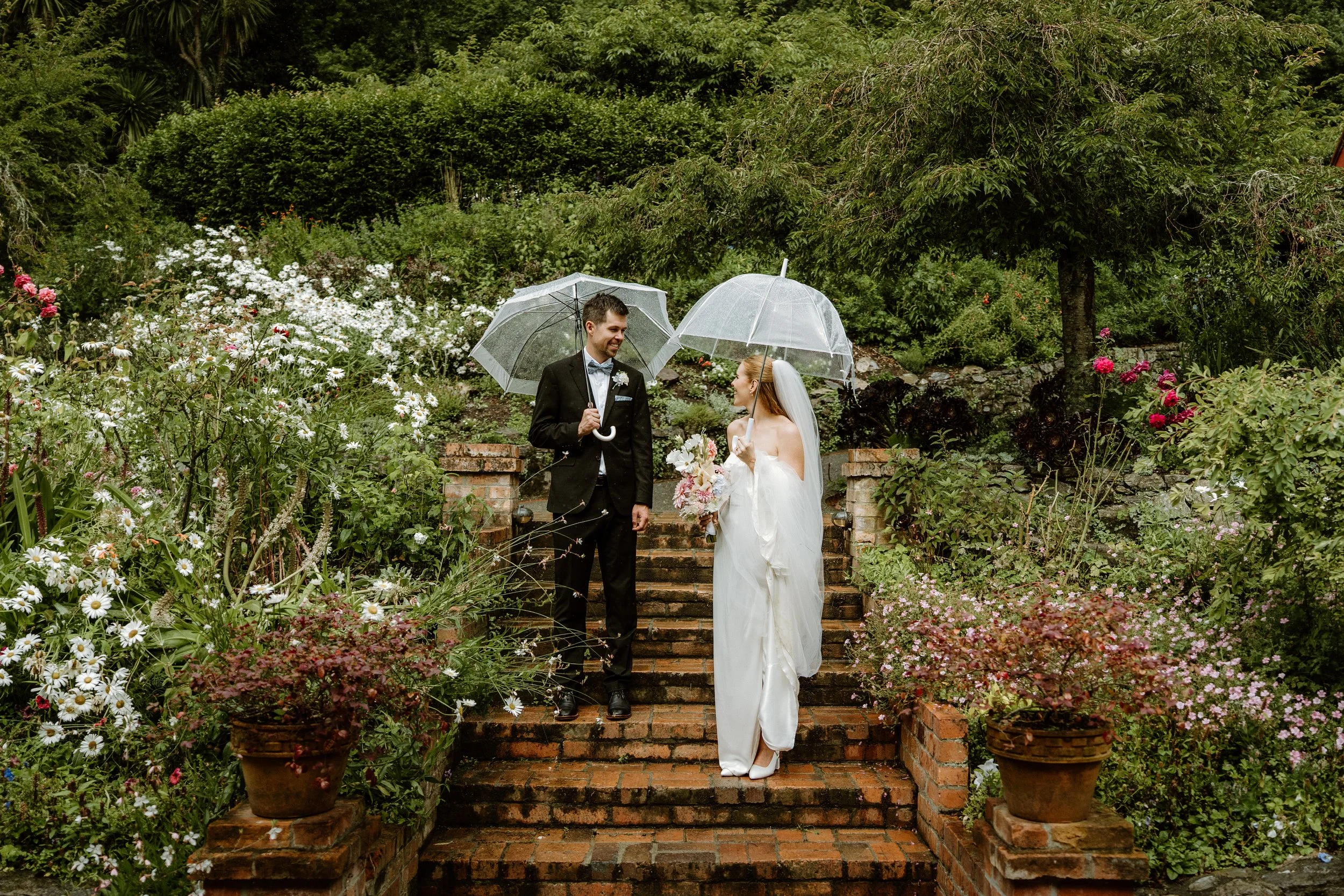 A bride and groom holding umbrellas standing on brick steps in a lush garden during a wedding.