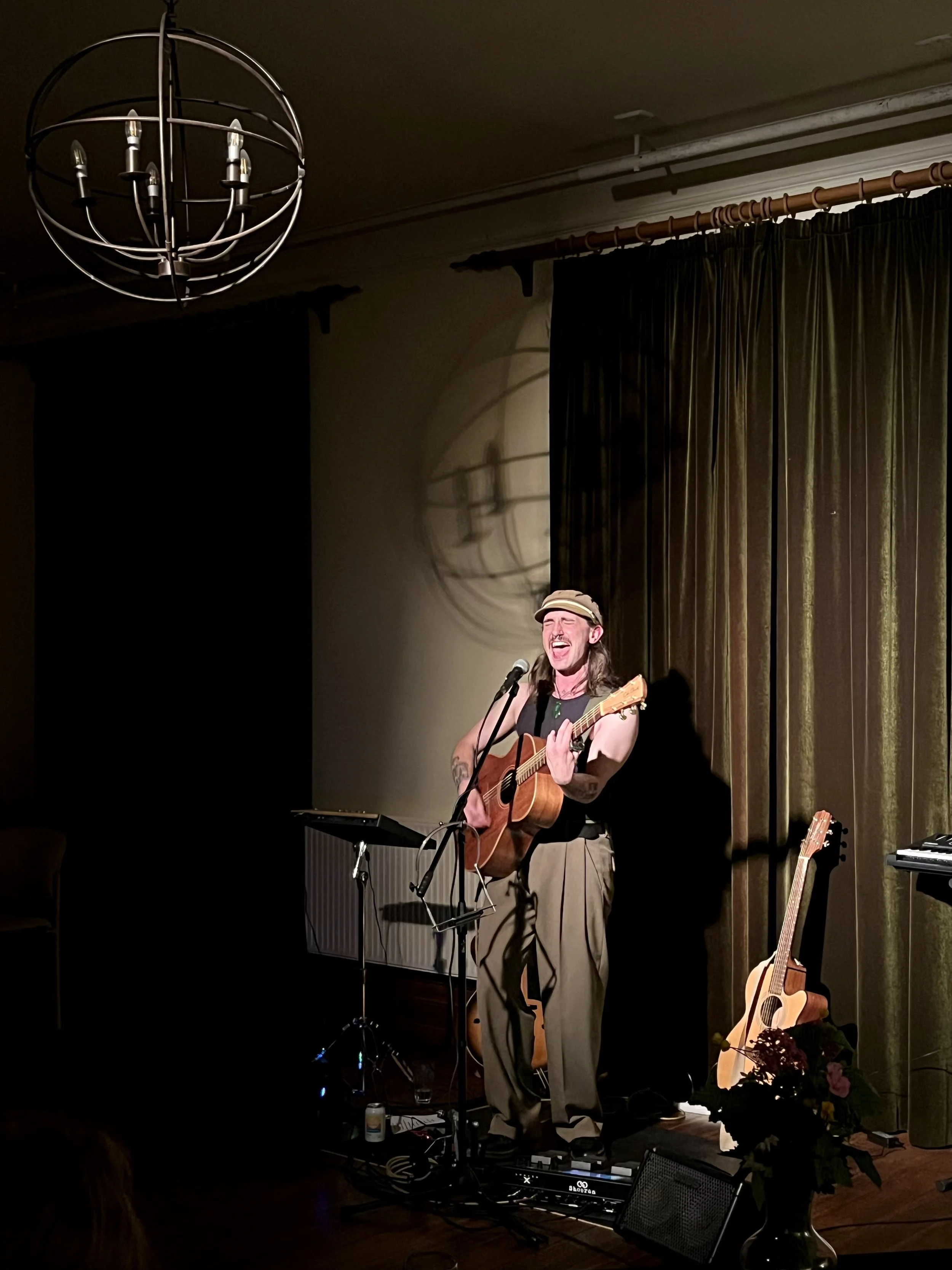 A man singing and playing an acoustic guitar on stage with a microphone in front of him, illuminated by warm stage lighting, with a floral arrangement and another guitar on a stand nearby, and a chandelier hanging from the ceiling.