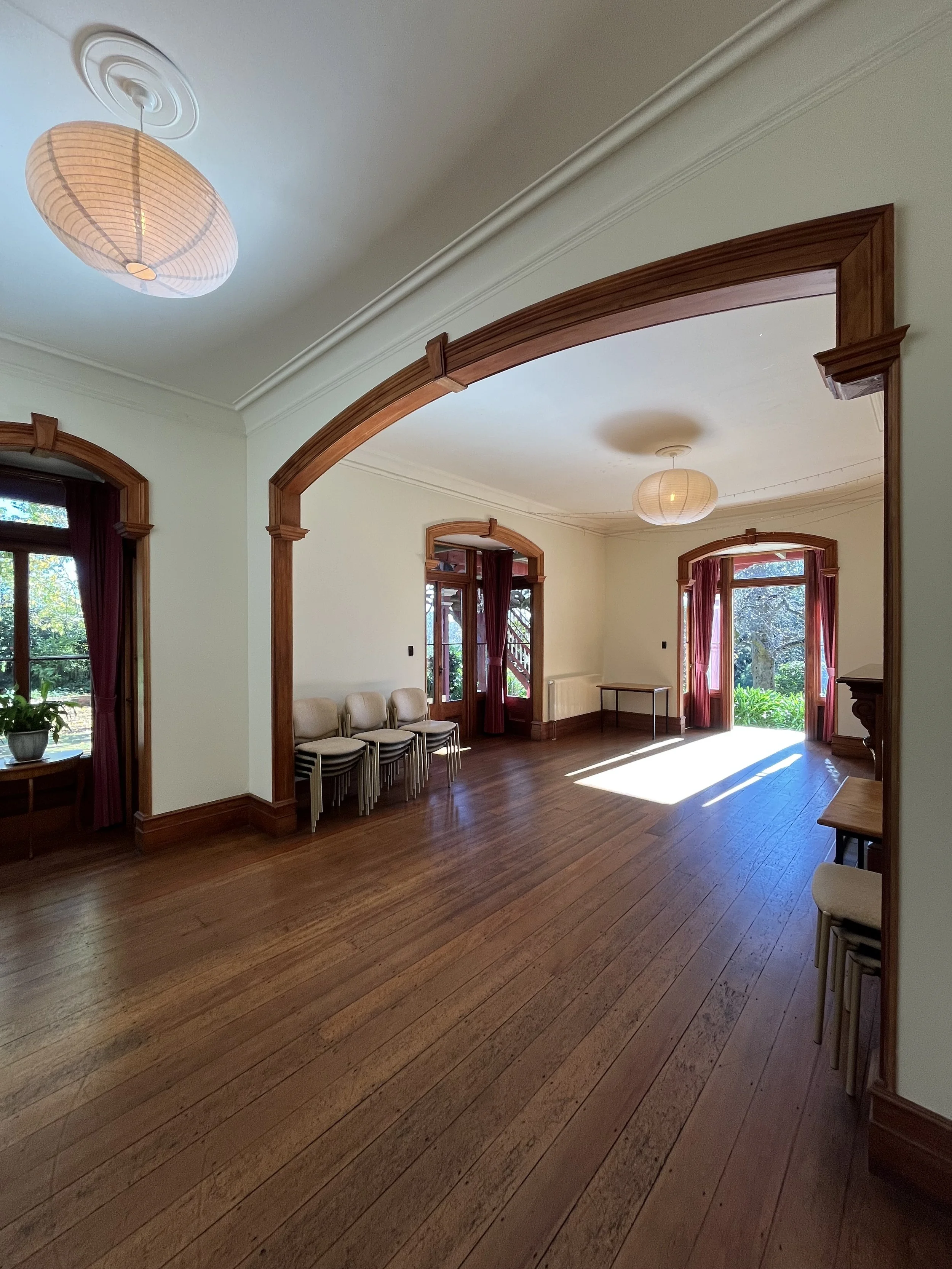 A room with large windows dressed with dark red curtains, wooden trim, hardwood floors, a few chairs stacked against the wall, and two ceiling lamps with paper shades, a small table near the windows, and a door leading outside.