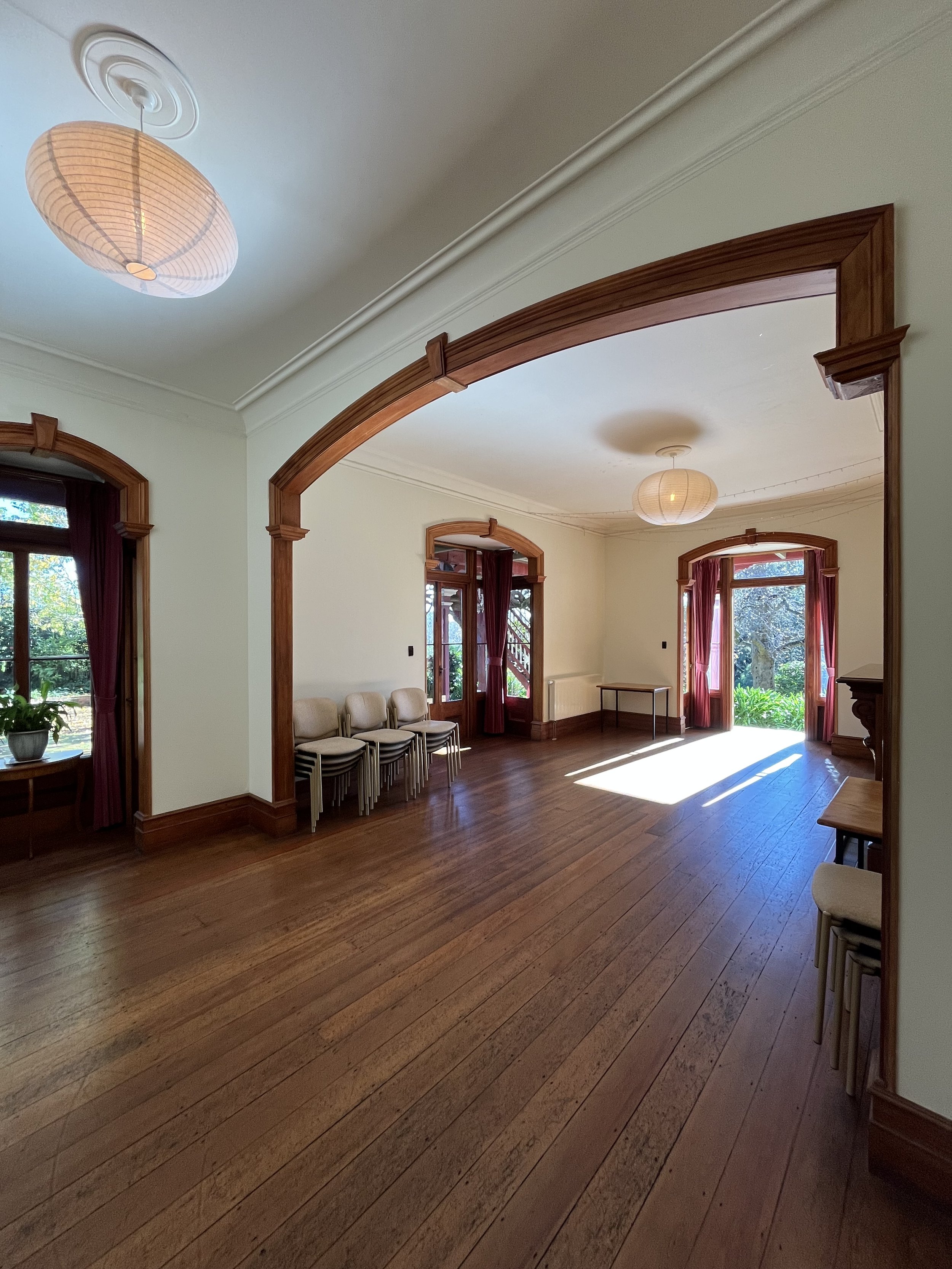 A spacious room with wooden floors, large windows with red curtains, and two ceiling lights. Several chairs are stacked along the walls, and a small table is near an open door that leads outside to a garden.