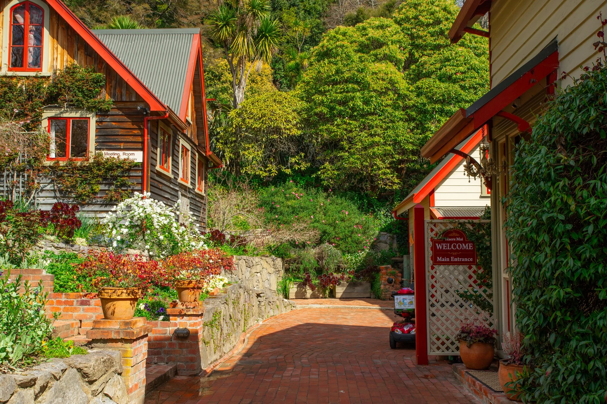 A charming garden pathway with brick pavers, flanked by lush greenery and colorful flowers, leads to a wooden building with a welcoming sign.