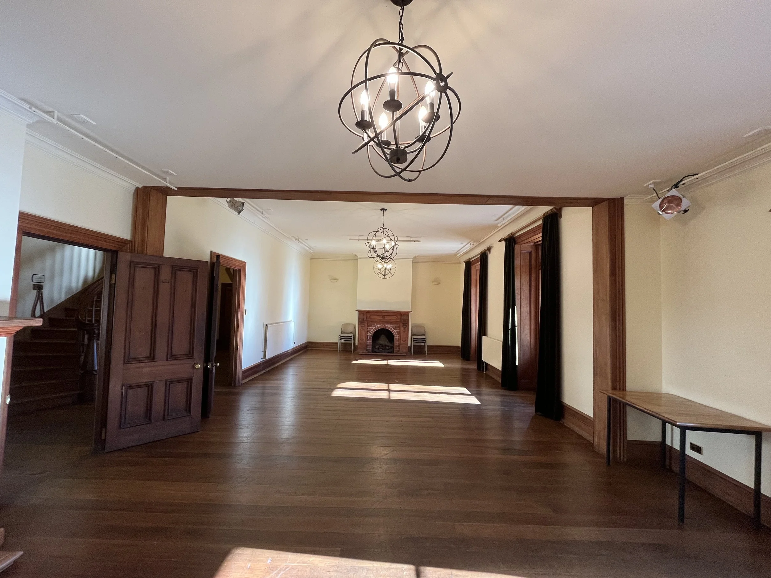 Large empty room with wood floors, beige walls, high windows with dark curtains, a decorative chandelier hanging from the ceiling, and a brick fireplace with a wooden mantel.