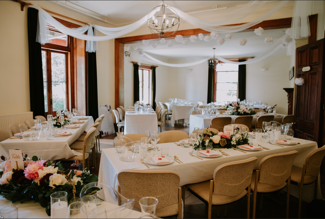 A decorated banquet room with tables set for a wedding reception or similar event, featuring floral centerpieces, white tablecloths, elegant glassware, and draped white fabric and paper pom-poms hanging from the ceiling.
