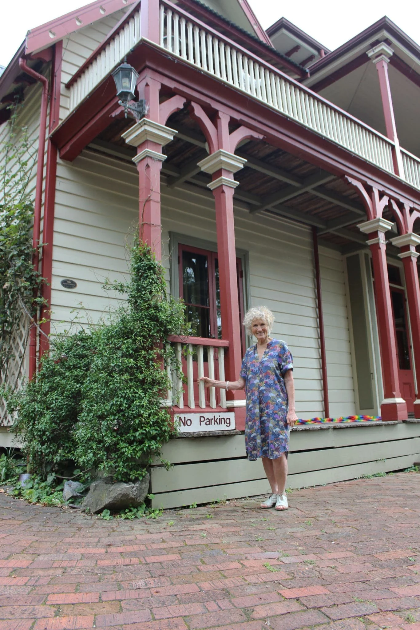 An elderly woman with curly white hair standing on a brick sidewalk in front of a two-story Victorian house with cream siding and red trim. She is smiling, wearing a colorful floral dress with short sleeves and white shoes, holding a rainbow-colored object in her left hand. There is a bush with green leaves partially covering the house's front porch, which has decorative support beams. A small sign on the house reads "No Parking."