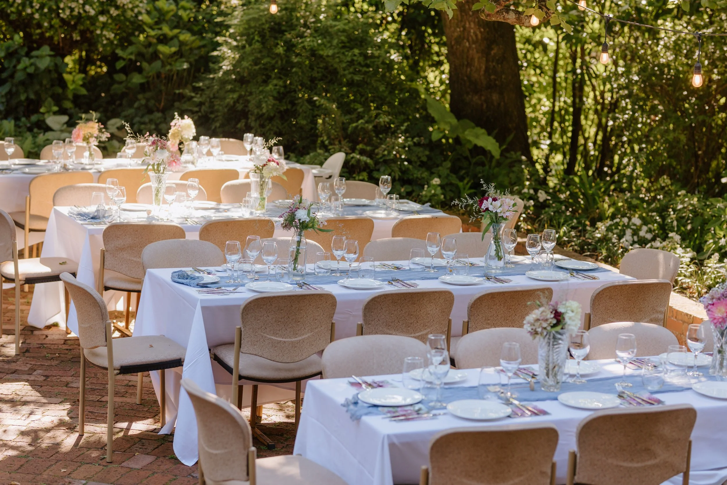 Outdoor dining setup with round tables covered in white tablecloths, decorated with flower arrangements and set with glasses, plates, and utensils, surrounded by beige chairs. String lights hang above in a lush, green garden setting.