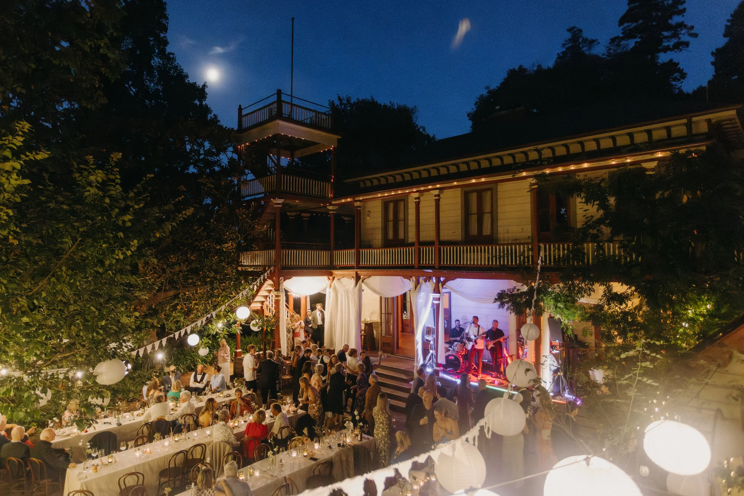 Outdoor evening celebration with people dining at long tables, a band playing on a stage, string lights, lanterns, trees, and a building with a balcony under a night sky with the moon.