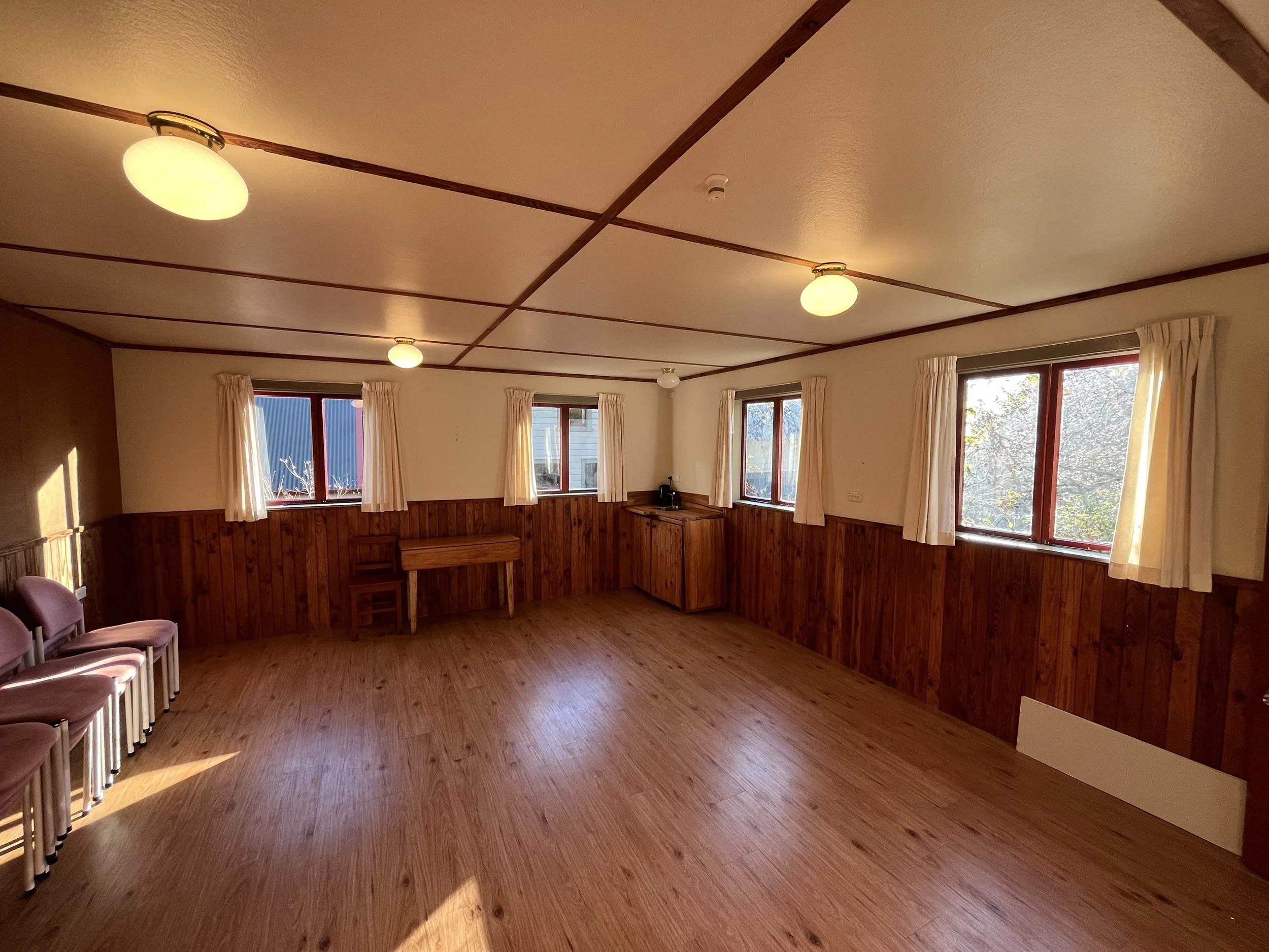 A spacious room with wooden flooring, beige curtains on windows, and ceiling lights.