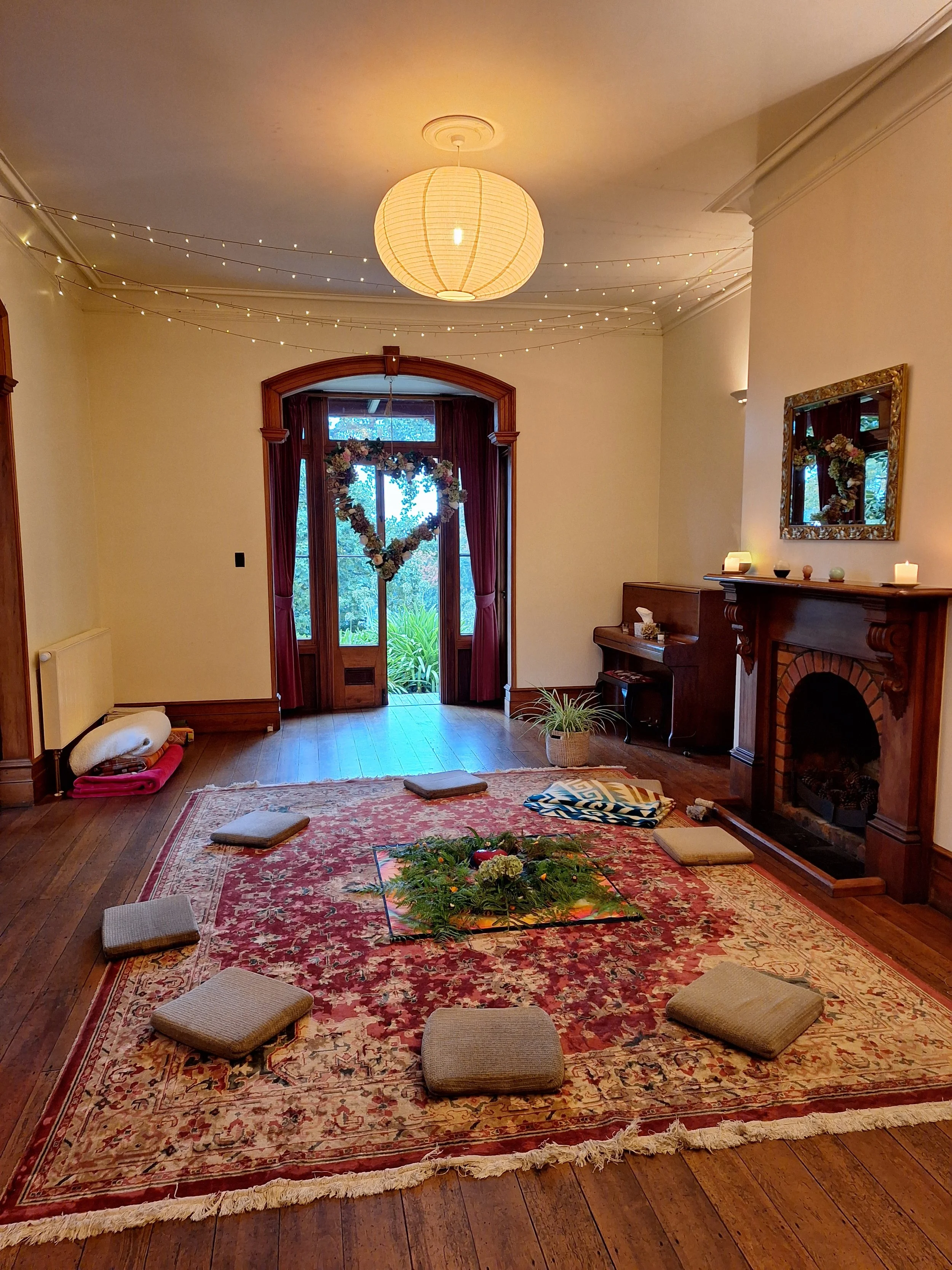 A cozy living room decorated for a special occasion with a large Persian rug, cushions on the floor, a potted plant, a fireplace with candles, a mirror, and a floral heart-shaped wreath in front of a window. String lights and a paper lantern hang fro