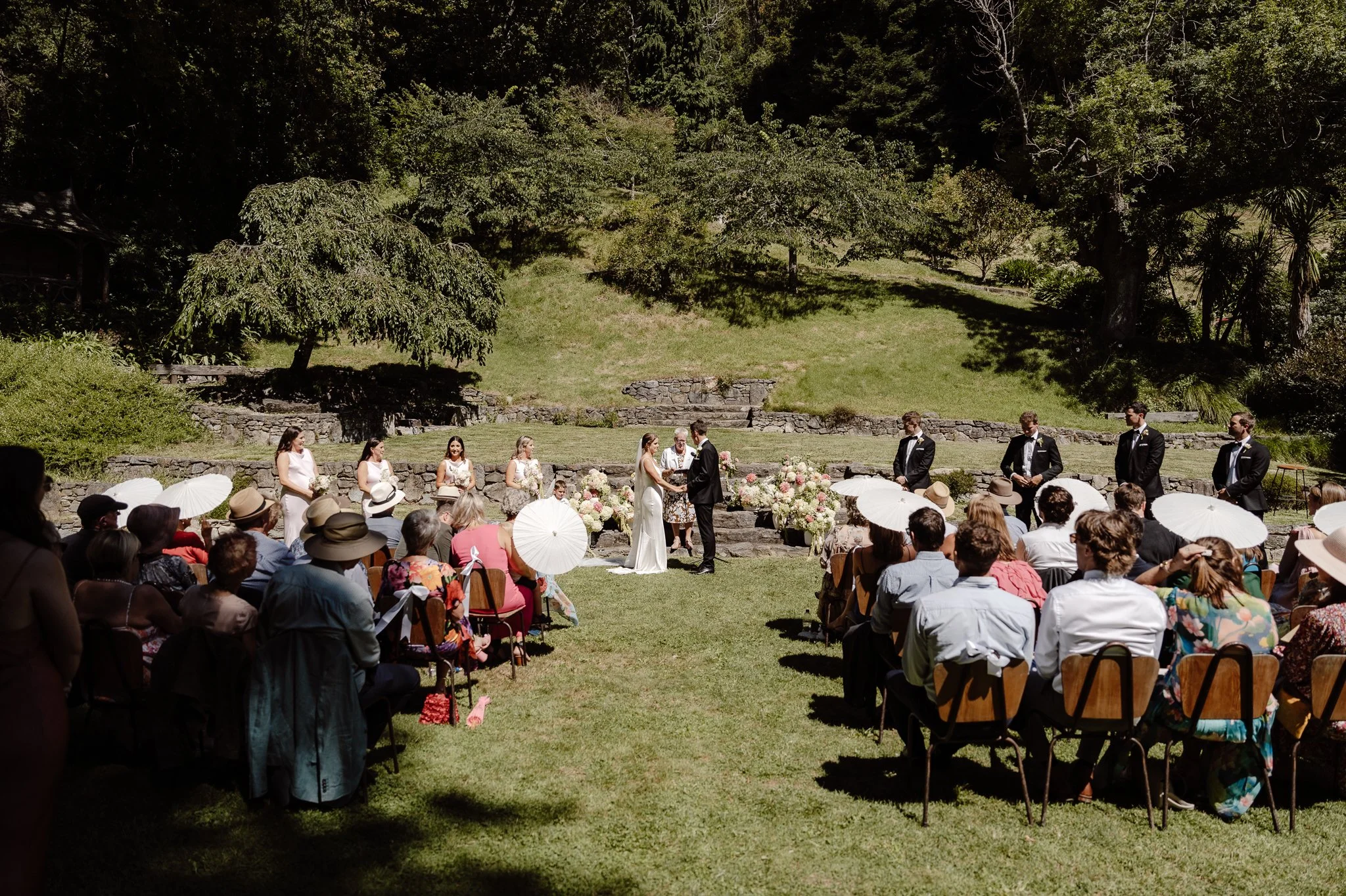 Outdoor wedding ceremony with guests seated on chairs, some holding umbrellas, in a lush green park with trees in the background. The bride and groom are exchanging vows in front of an officiant, with bridesmaids and groomsmen standing nearby.