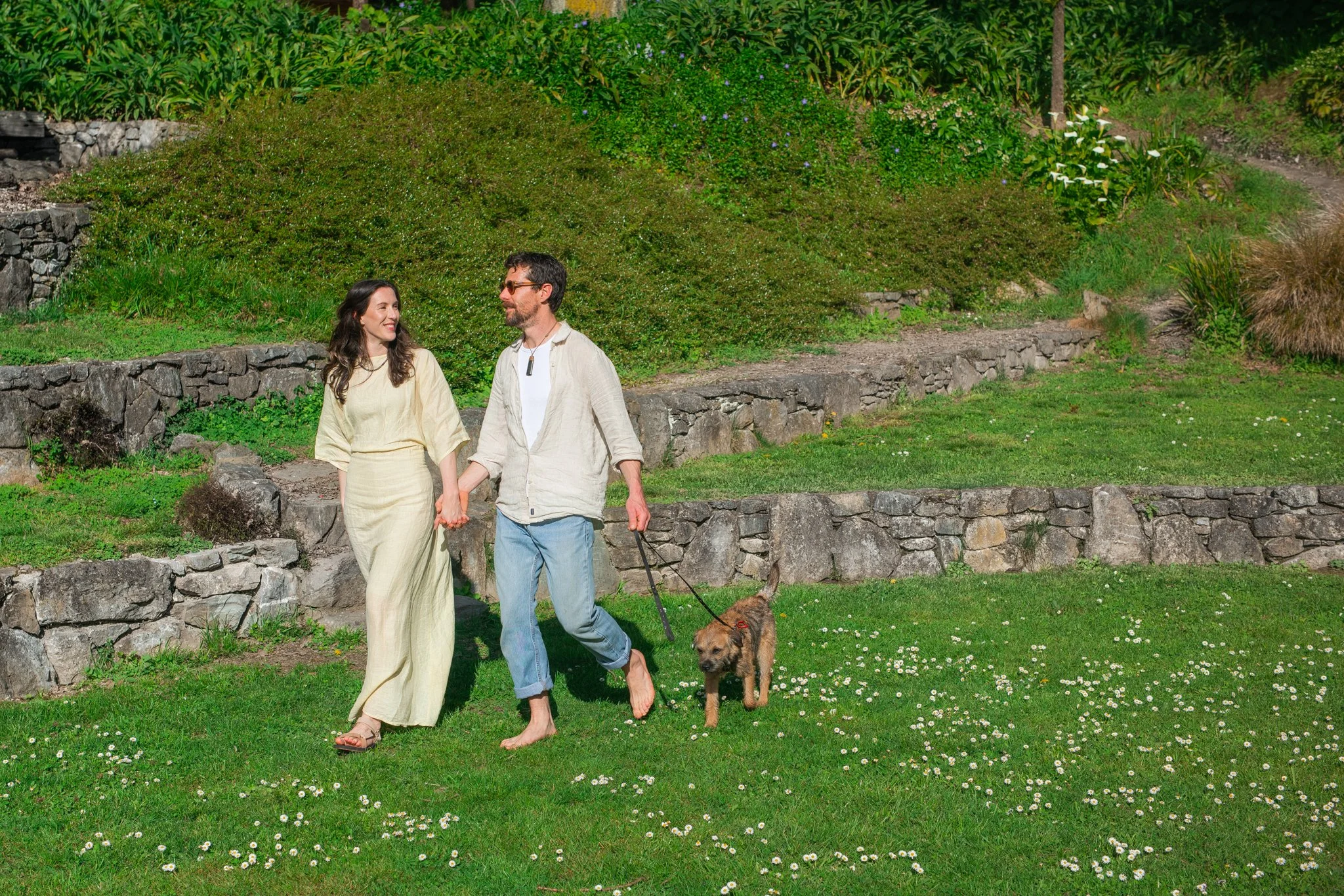 A woman and a man walking hand in hand with two dogs on a grassy area with white flowers, surrounded by greenery and stone steps.