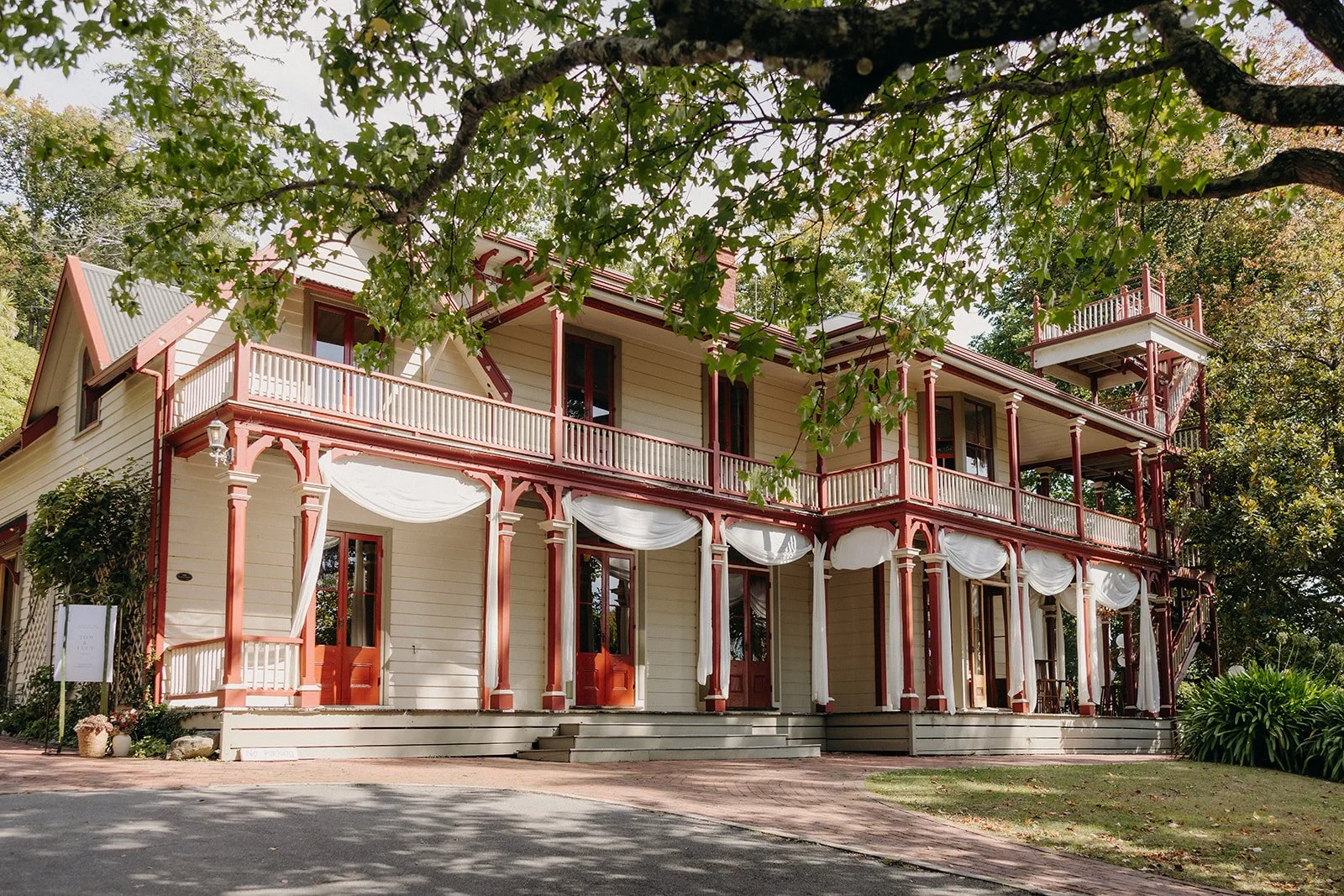 A large, three-story Victorian-style house with cream-colored siding and red trim. It has a wraparound porch with white drapes and a staircase leading up to the front door, surrounded by trees and greenery.