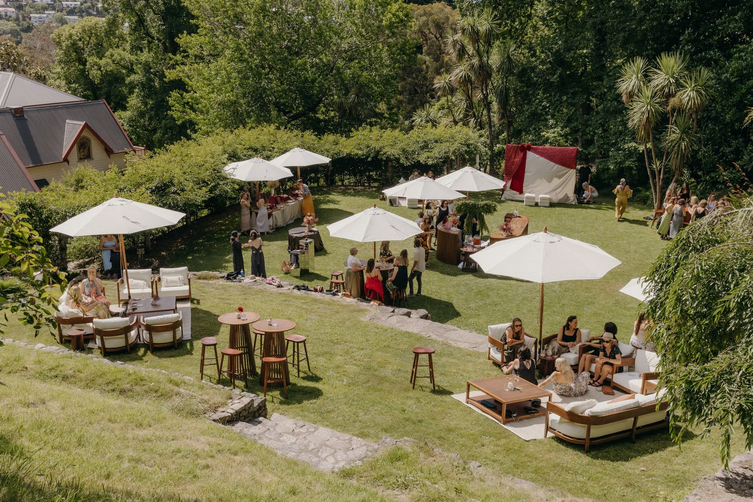 An outdoor gathering with people socializing on a grassy lawn surrounded by trees. There are white umbrellas, seating areas, a red and white stage, and tables with food and drinks. Some people are standing, others seated, and a few are dancing or talking.