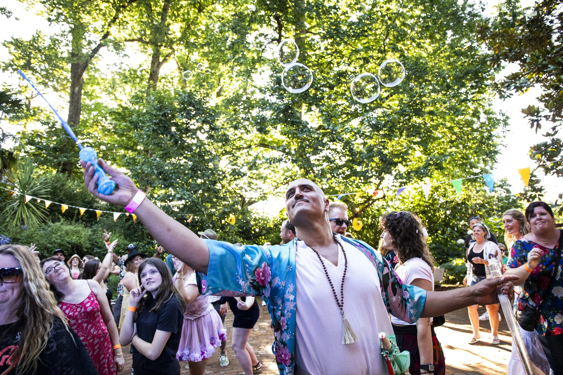 A diverse group of people enjoying an outdoor festival or celebration in a lush, green setting, with some blowing bubbles and others dancing or watching.