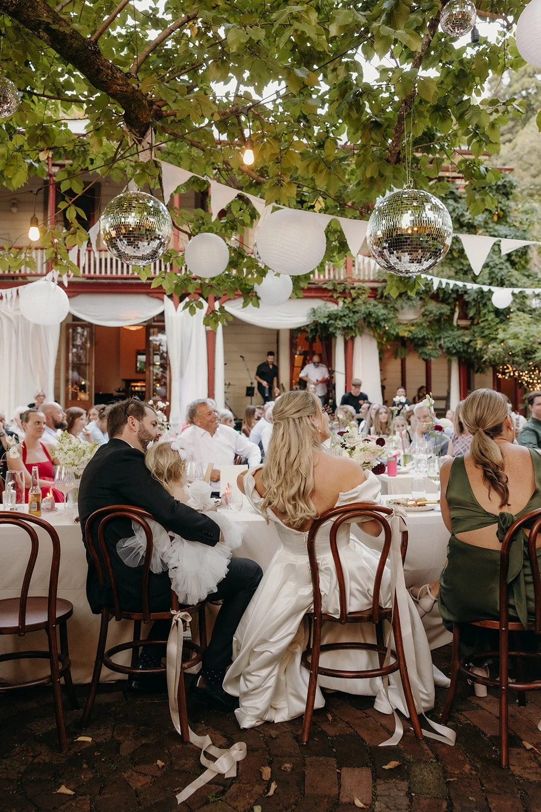 A wedding reception outdoors with guests seated at decorated tables under trees with hanging lanterns and disco balls, and a live band playing on a stage in the background.