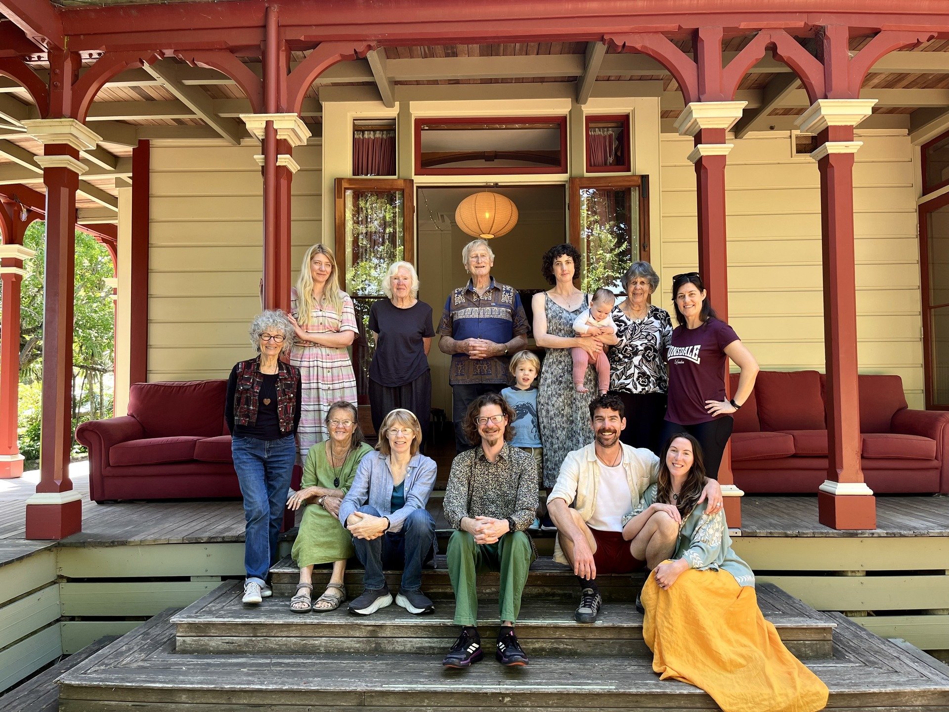 Group of diverse people gathered on the porch of a yellow house with red trim, sitting and standing, smiling, some children present at daytime.