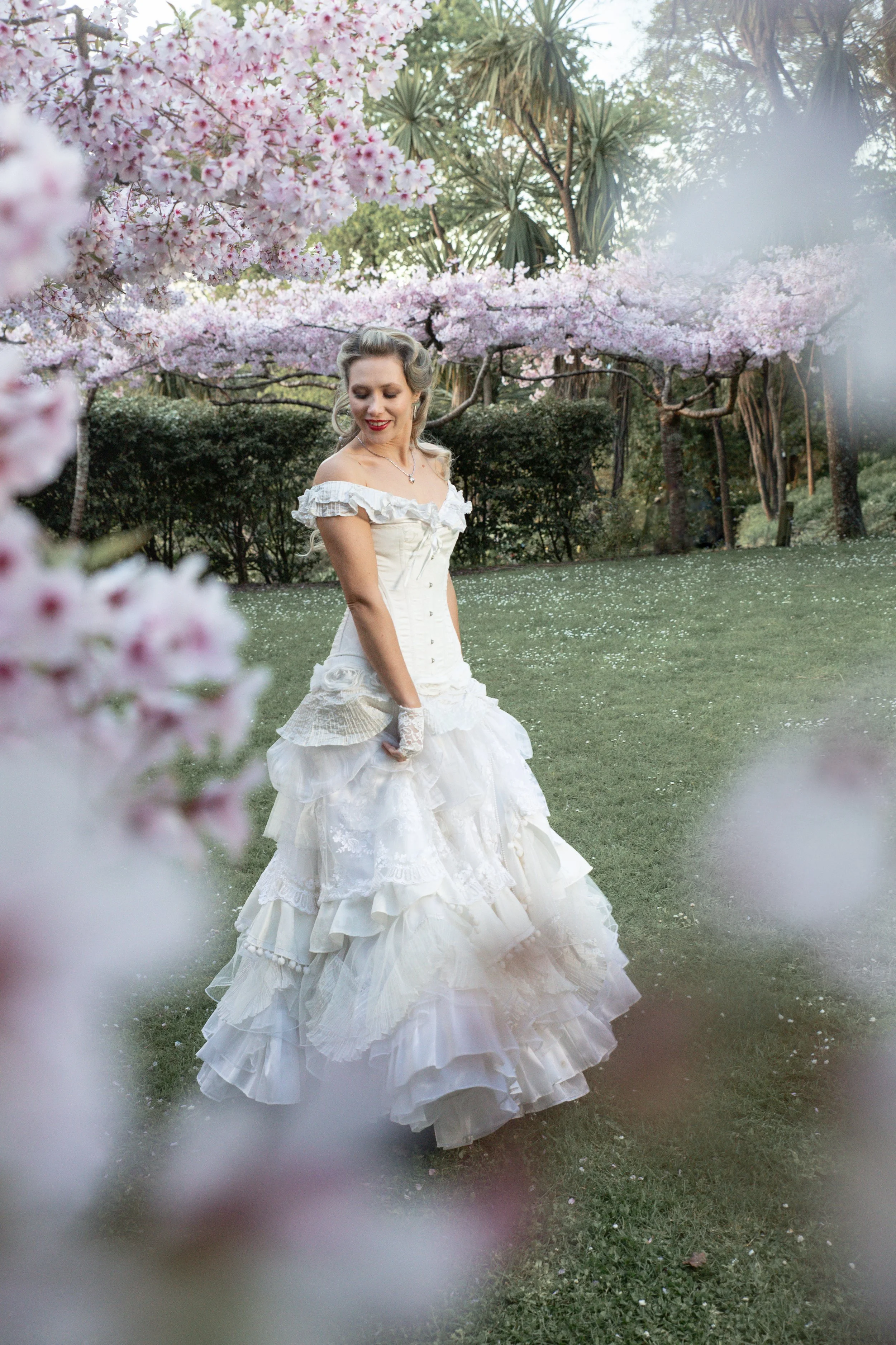A woman in a vintage white wedding dress with ruffled layers and off-the-shoulder neckline is standing outdoors in a garden with pink cherry blossom trees in full bloom and palm trees in the background. The woman is smiling and looking downward.