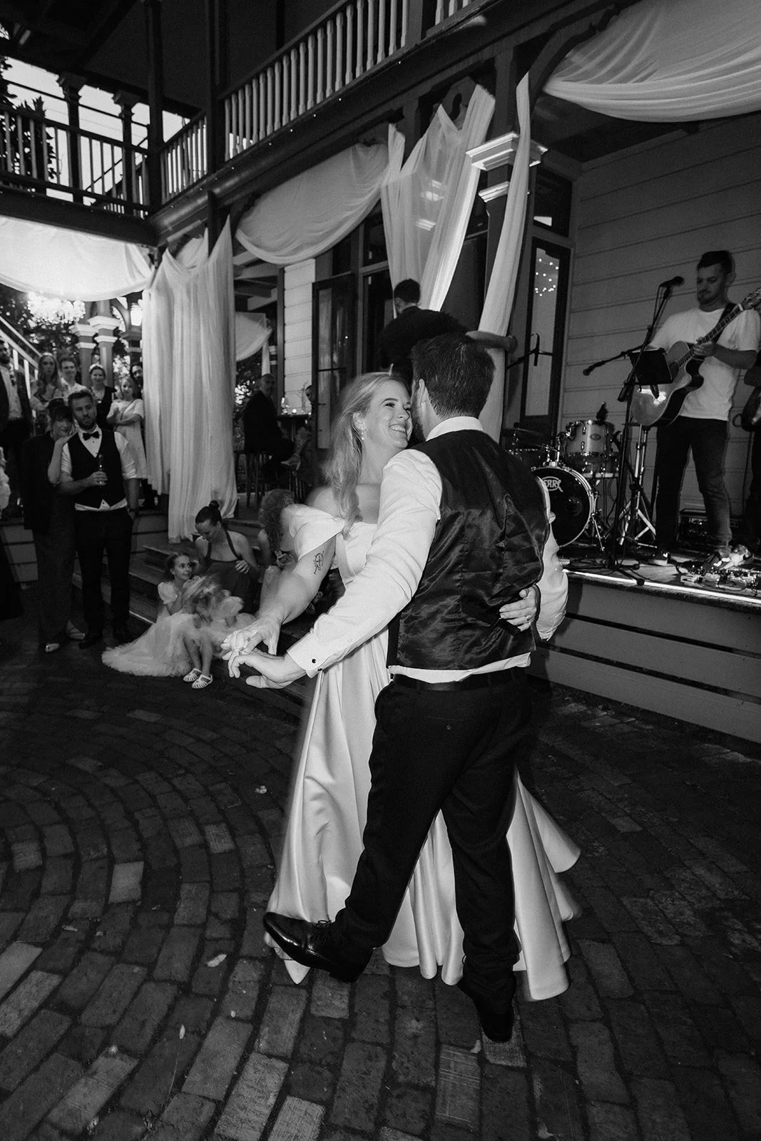A bride and groom dancing at their wedding reception, surrounded by guests and a live band, in an outdoor setting with draped fabric decor.