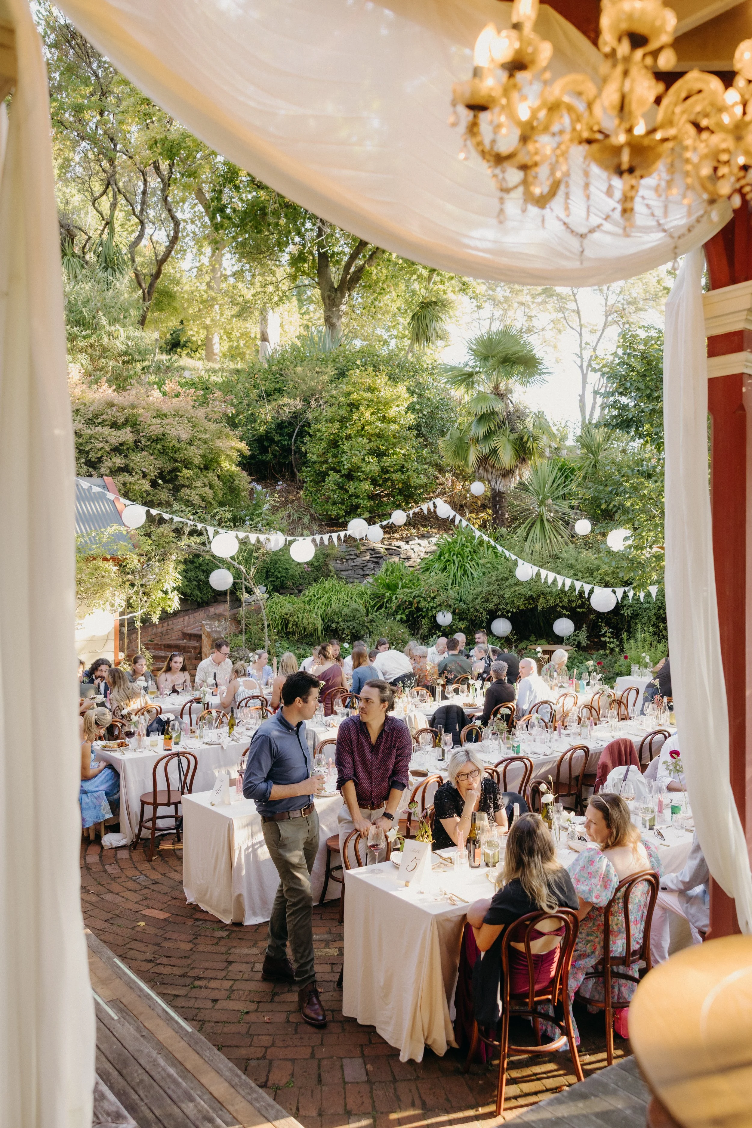 A lively outdoor wedding reception under a tent with white drapes, large flower arrangement, and a chandelier. Guests are seated at decorated tables, with some standing and talking. The background shows lush greenery, trees, and string lights.