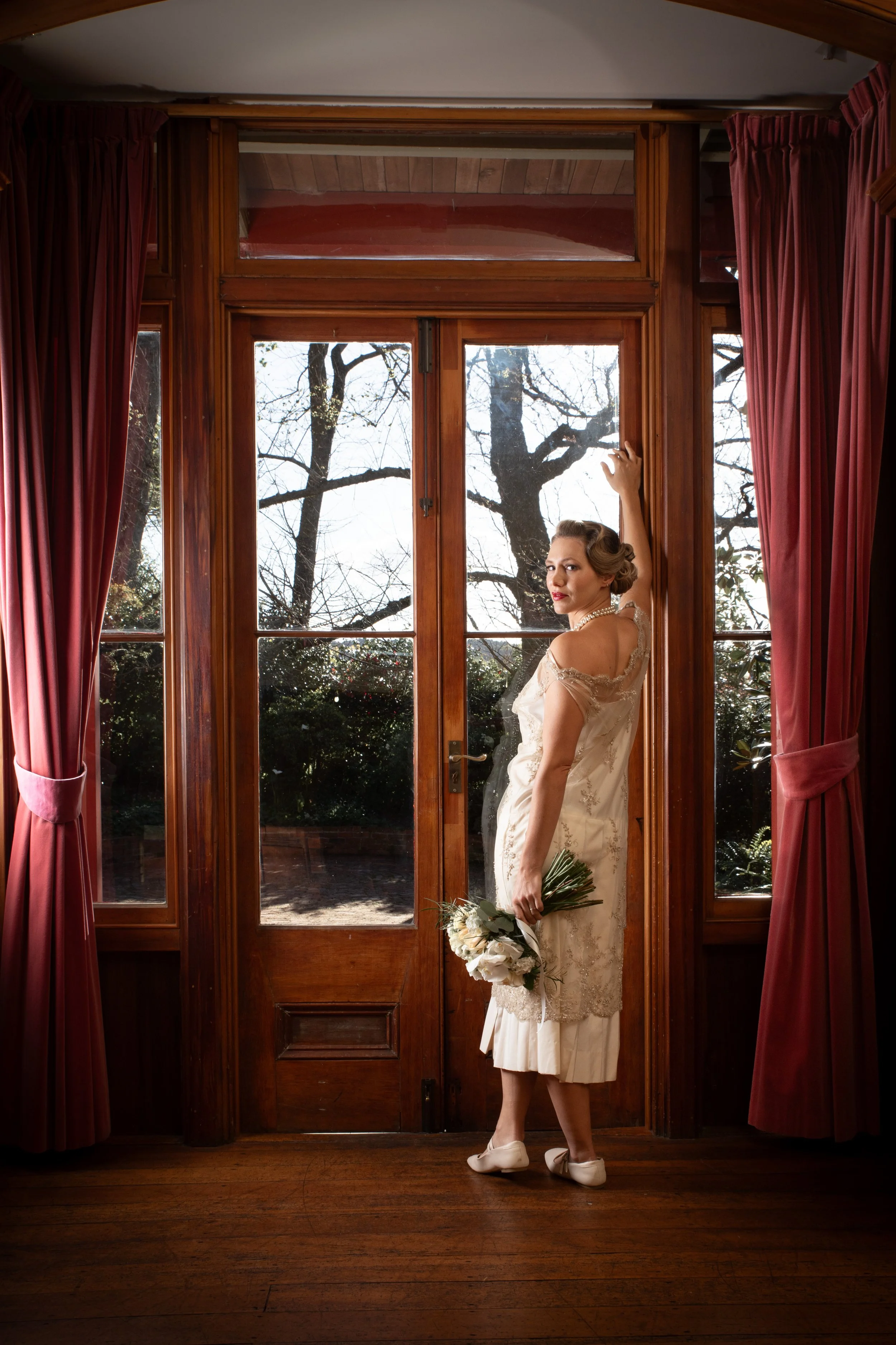 A woman in a vintage wedding dress stands in front of a wooden door with glass panes, holding a bouquet of white flowers, with trees visible outdoors through the glass.