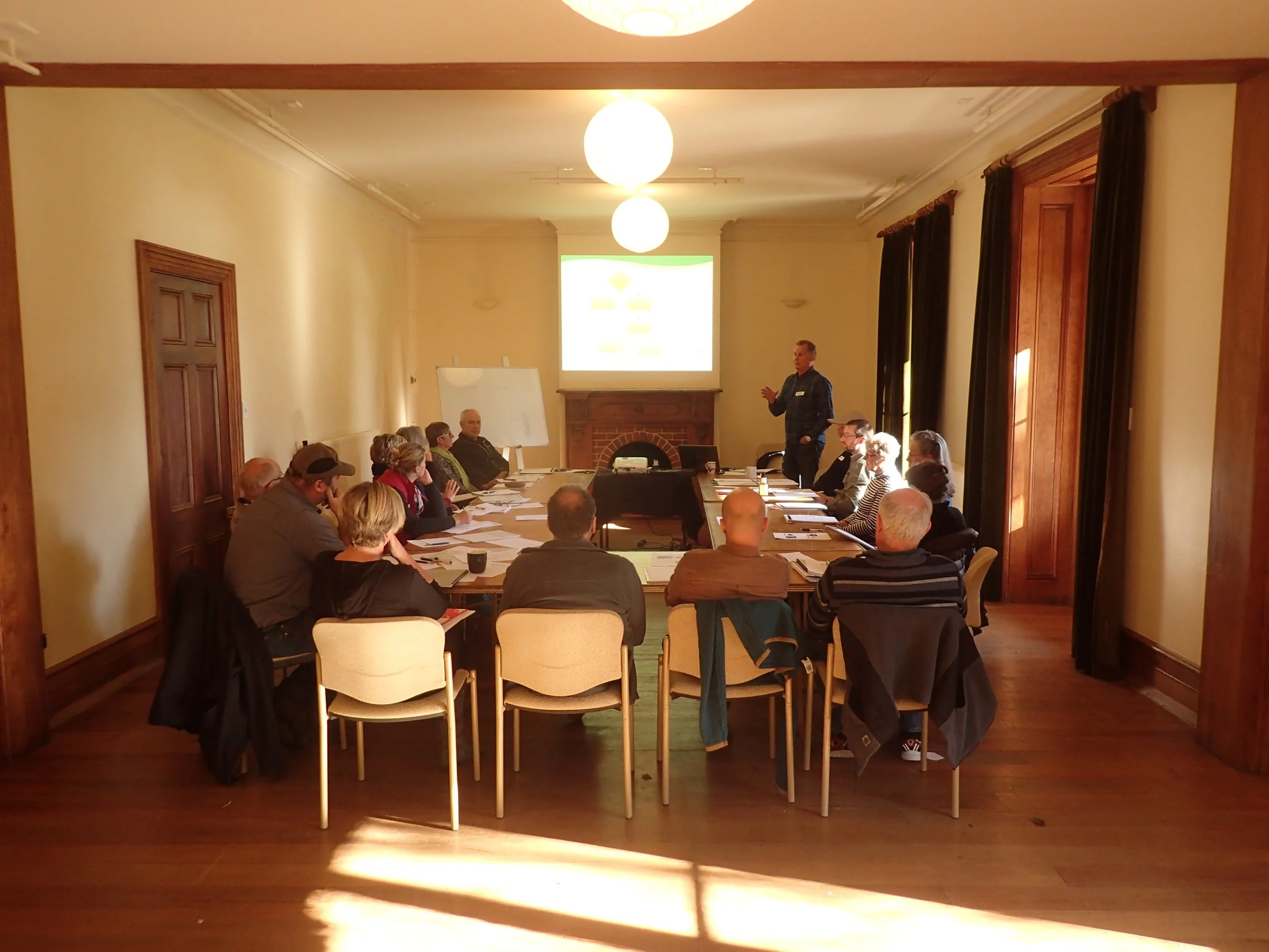 A group of people seated around a large table in a conference room, with a man standing and speaking near a whiteboard and a projection screen at the front of the room, and sunlight streaming in through the windows covered with dark curtains.