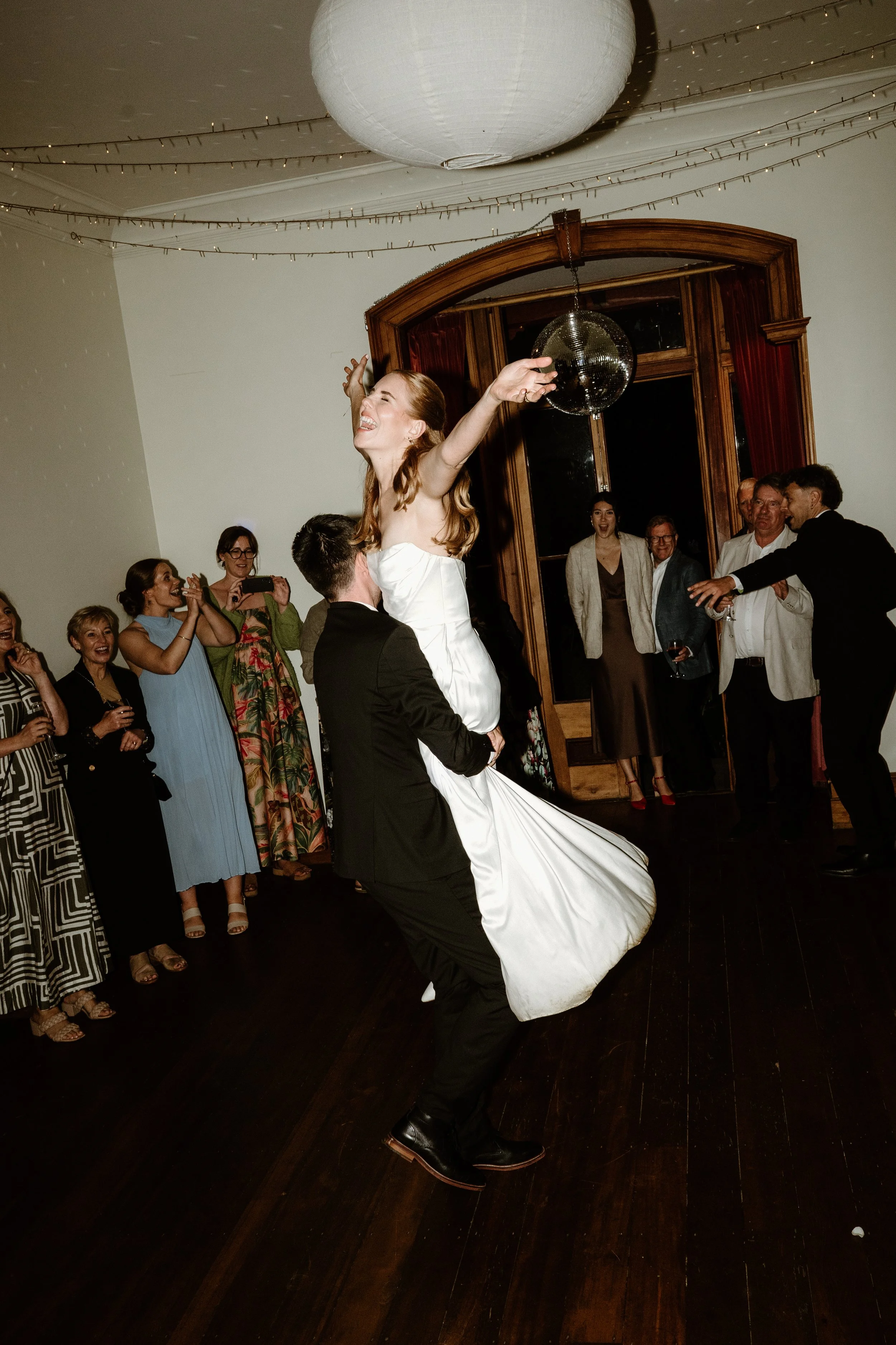 A wedding reception with a bride and groom dancing as guests watch and take photos. The bride is being lifted by the groom and has her arms outstretched, wearing a white wedding dress. Guests are smiling, clapping, and capturing the moment in a room 