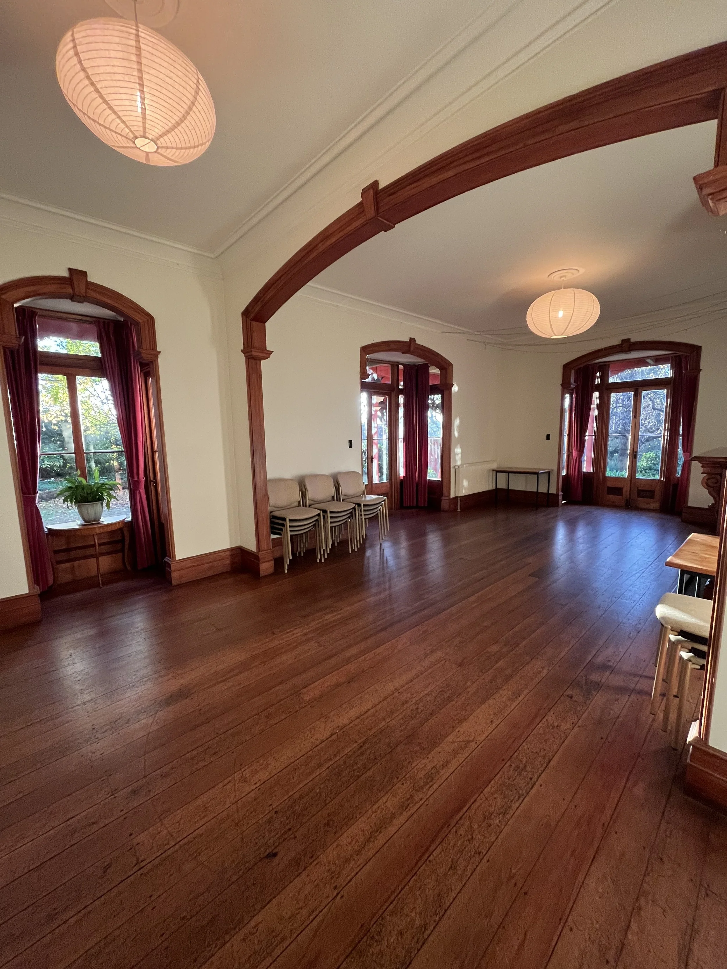 Empty room with wooden floors, large windows with red curtains, chairs stacked against the wall, and hanging paper lantern lights.