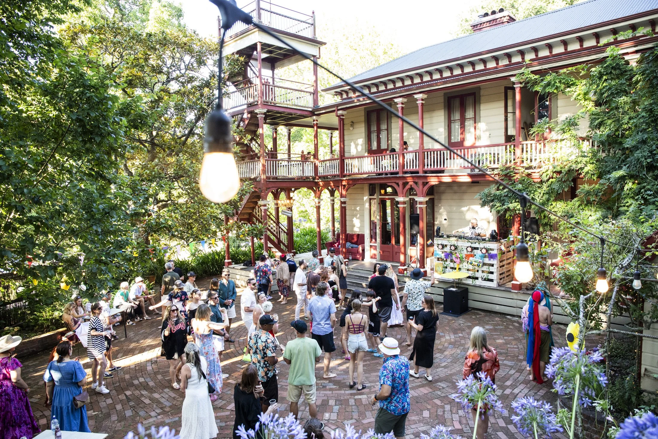 A lively outdoor gathering with people dancing and socializing in front of a house with a wraparound porch, surrounded by trees and string lights.