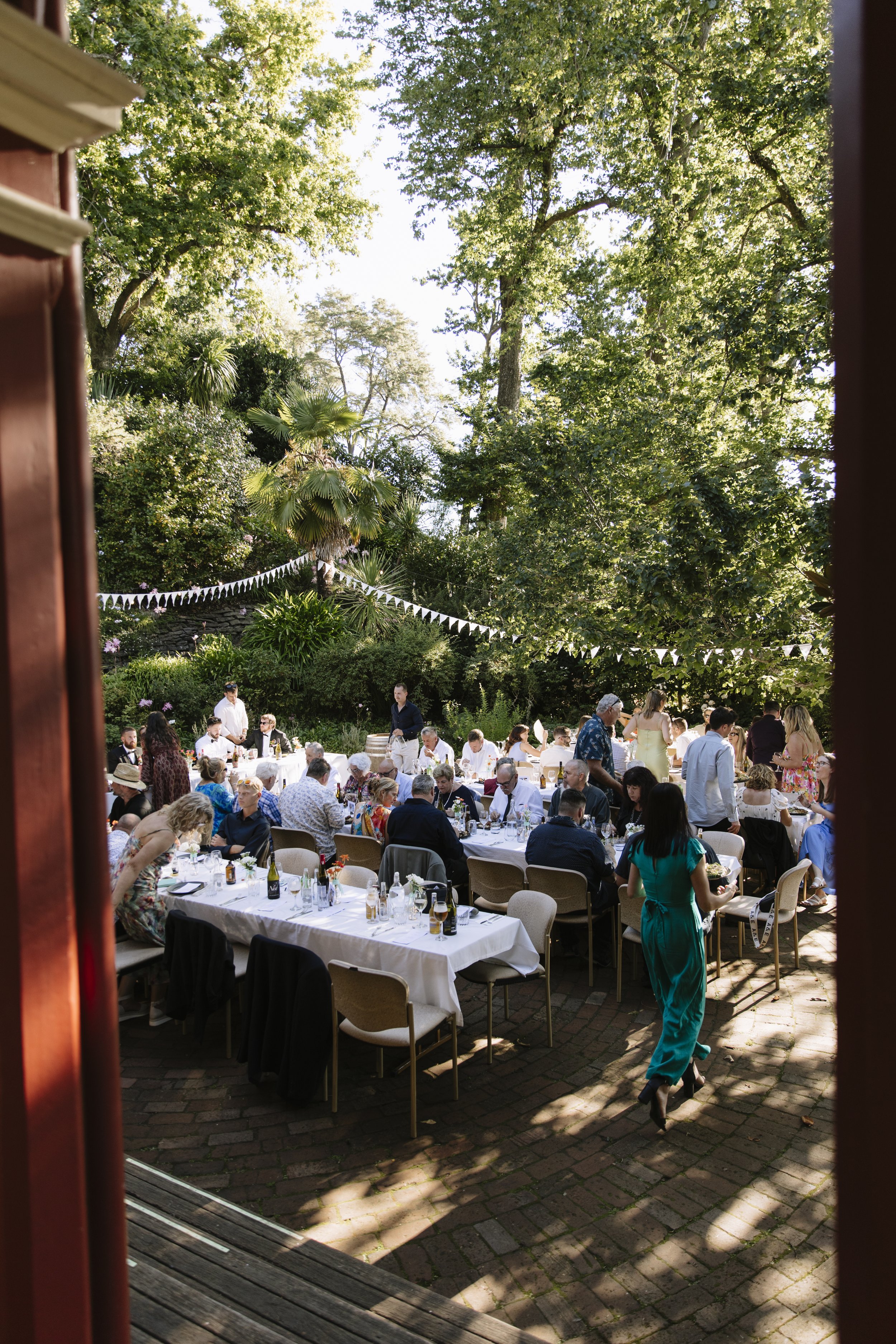 Outdoor celebration with many people seated at tables, under large green trees, with a woman in teal walking by. The scene is viewed through a doorway with red framing.
