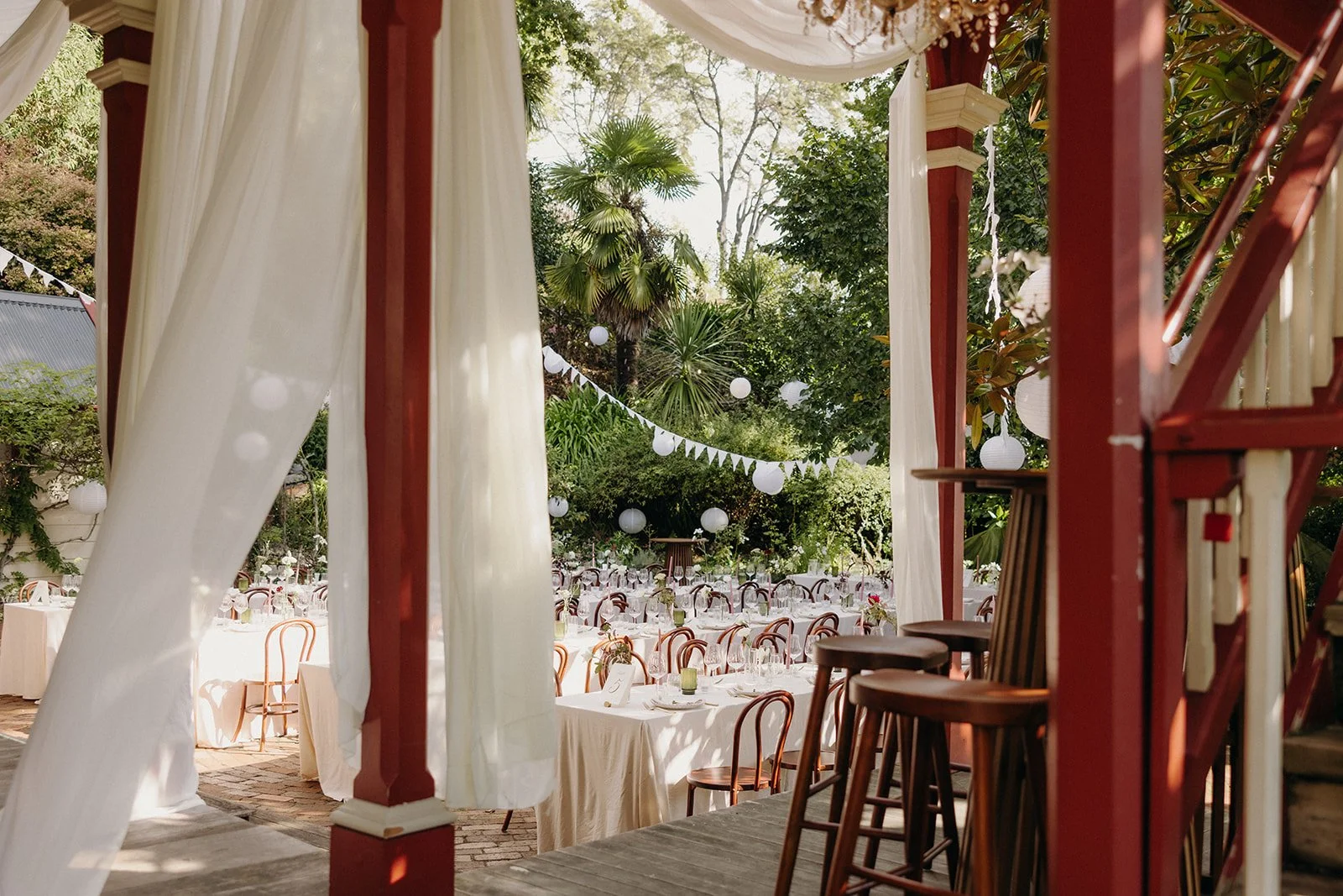 Outdoor event space decorated with white drapes, string lights, and tables set with glasses and centerpieces, surrounded by greenery and trees.