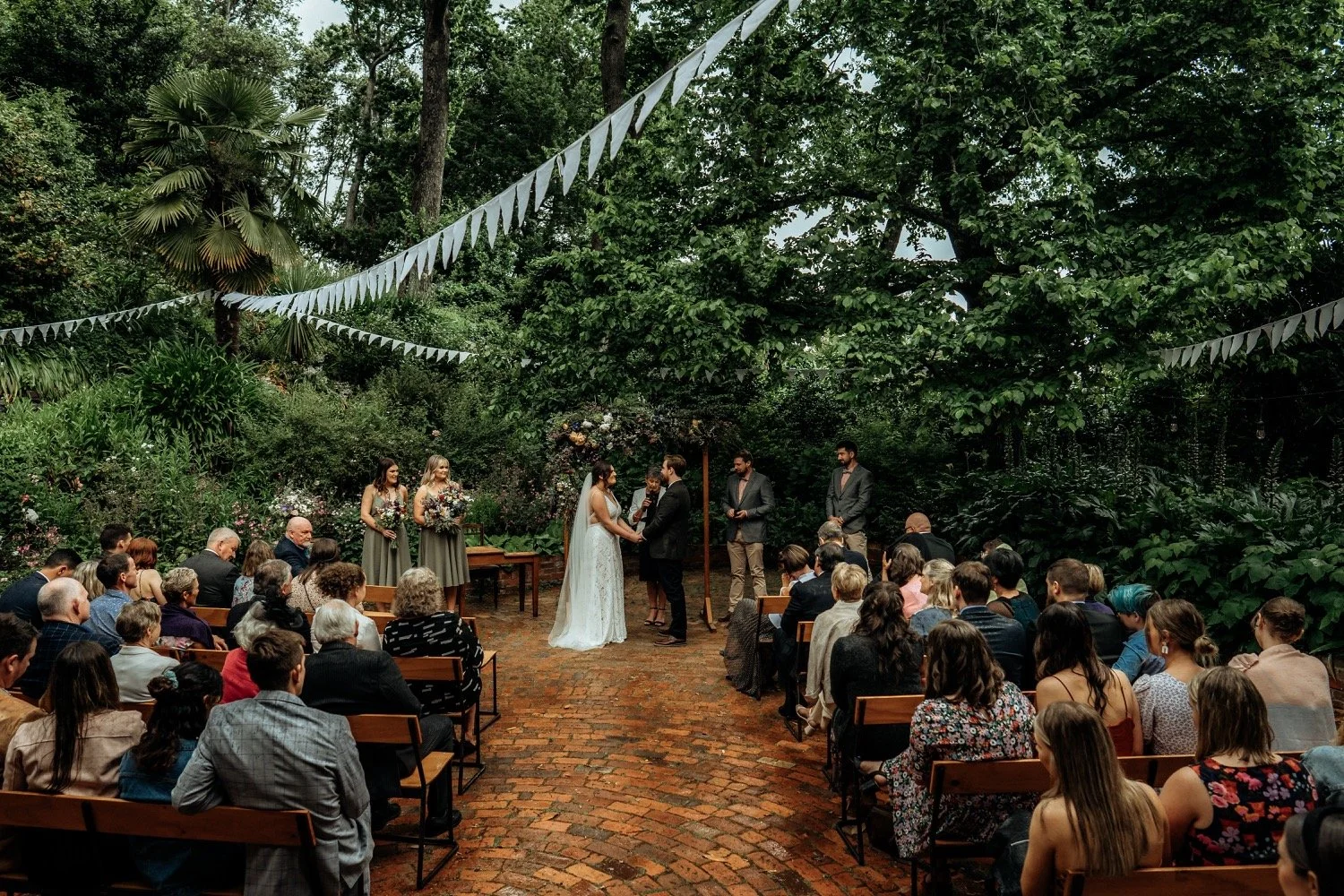 An outdoor wedding ceremony in a lush green garden with a brick pathway. A bride and groom exchange vows in front of an officiant, surrounded by bridesmaids, groomsmen, and seated guests. White bunting decorations hang above.