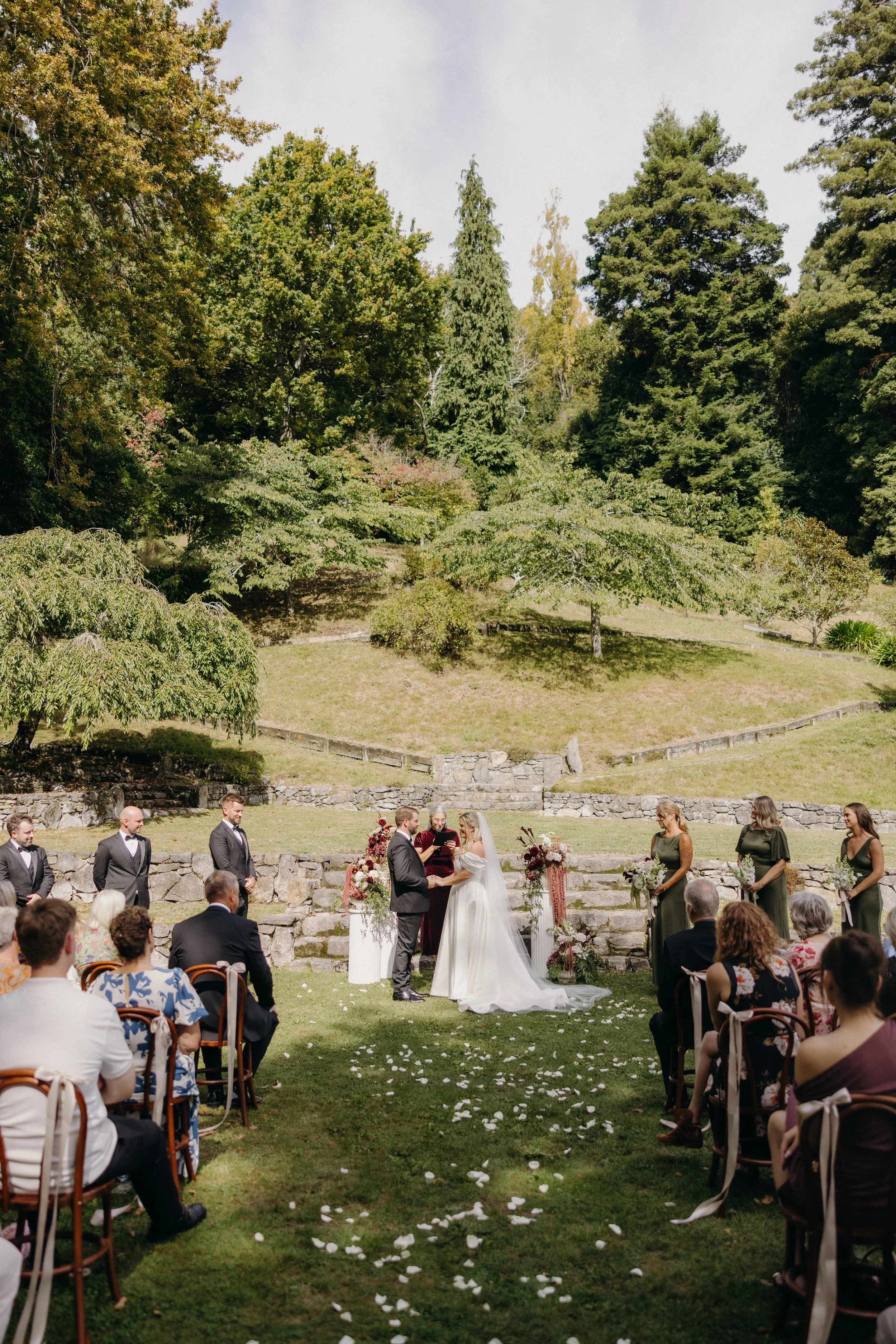 Outdoor wedding ceremony with a bride and groom exchanging vows, surrounded by wedding party and guests on a grassy area with stone steps and green trees in the background.