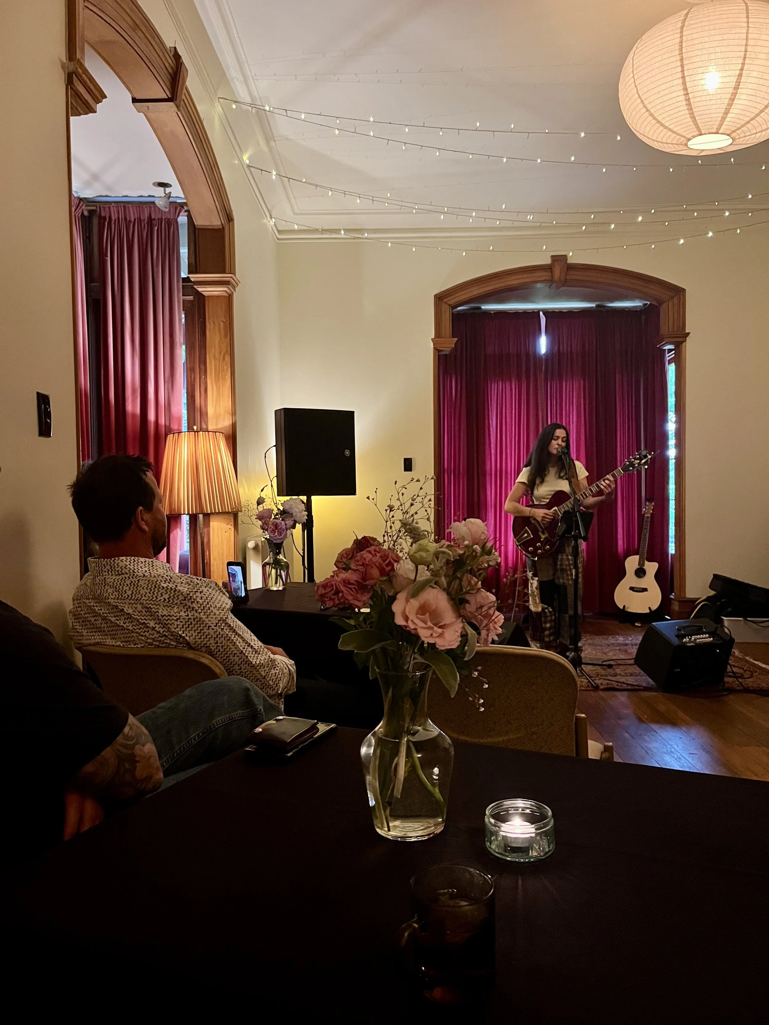 A woman playing guitar on a small stage with red curtains and string lights overhead, floral arrangements on the floor, and an audience seated at tables with flower centerpieces and candles.