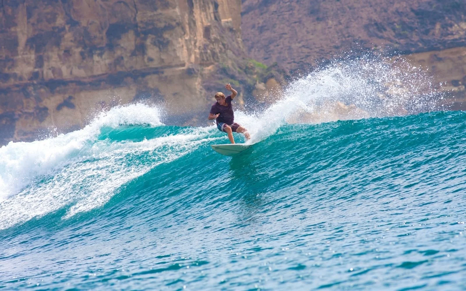 Person surfing on a blue ocean wave with a rocky cliff in the background.