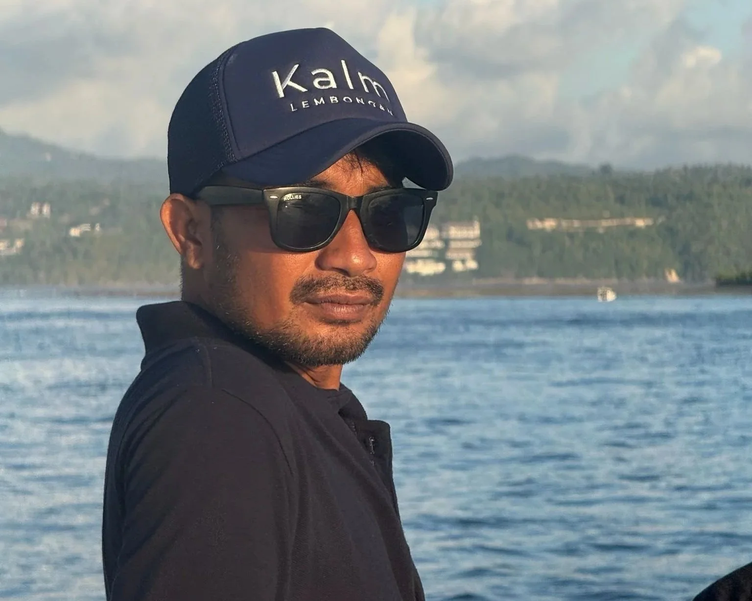 A man wearing a black cap with the words "Kalm Lemonade" and black sunglasses, standing outdoors near a body of water with hills and buildings in the background.