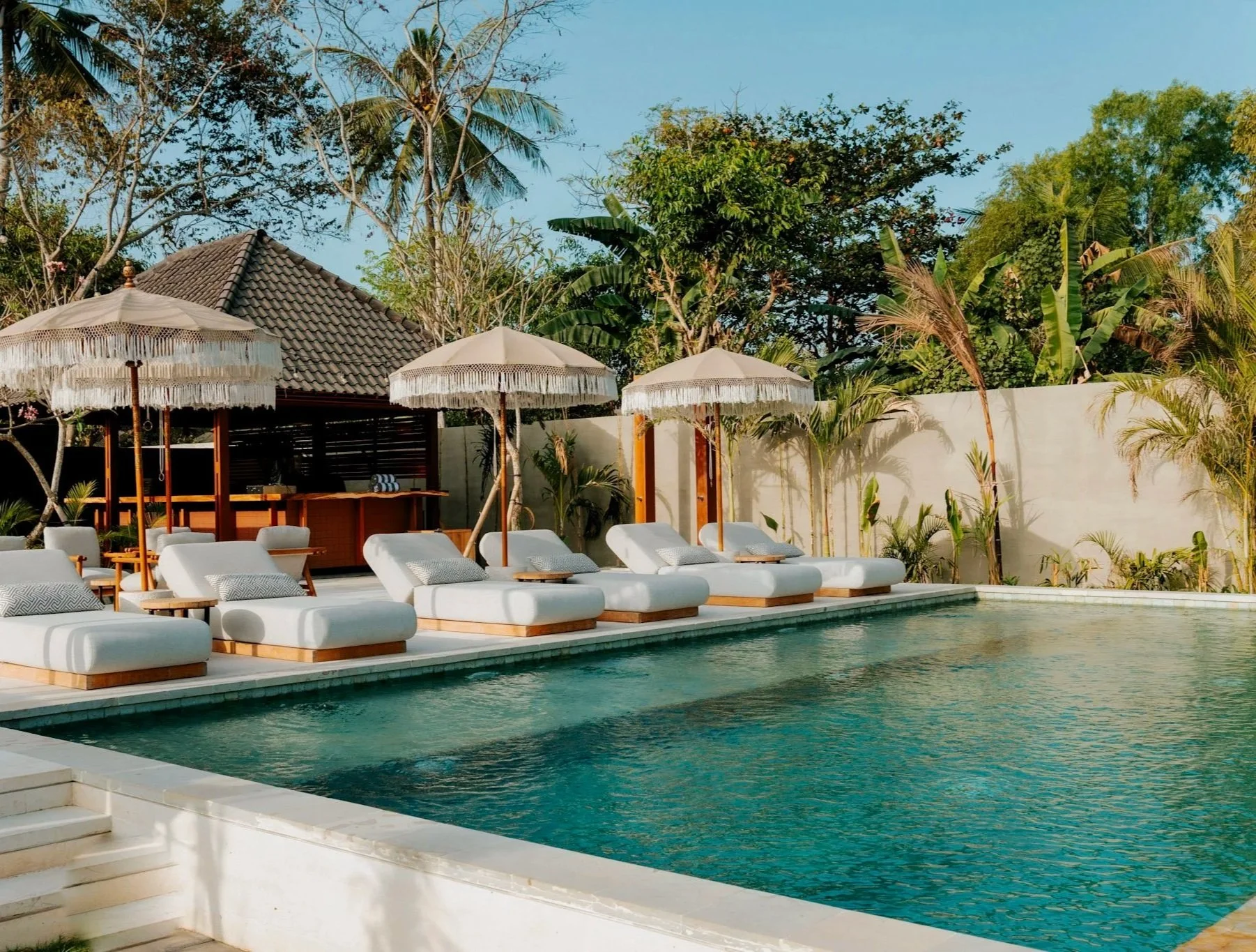 Luxury swimming pool area with white lounge chairs and umbrellas, surrounded by tropical trees and plants.