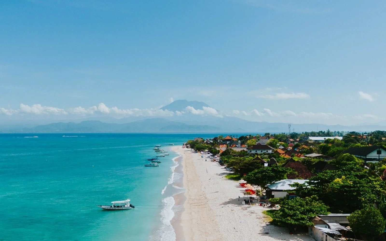 Tropical beach with blue water, boats, and a village with trees and houses in the background, and a mountain in the distance.