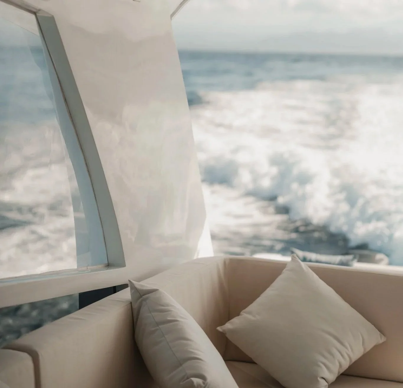 A view from inside a boat showing a window with white curtains and a comfy beige cushioned seating area with white pillows, overlooking the blue ocean with waves.