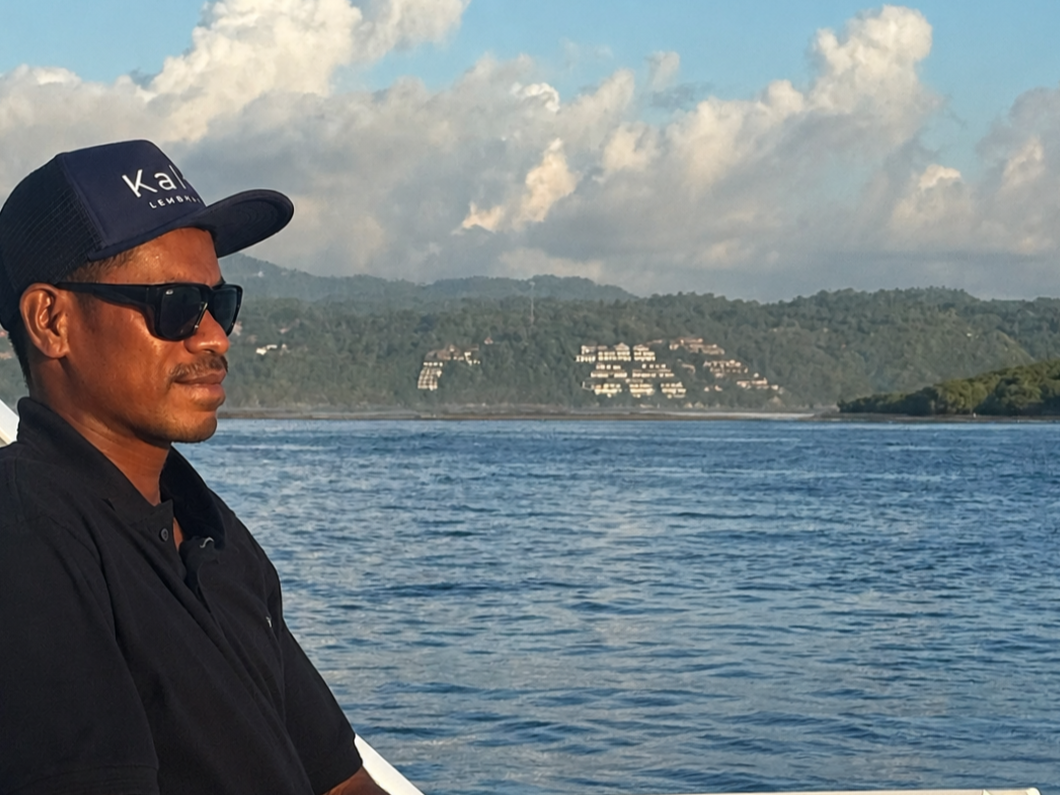 A man wearing sunglasses and a navy cap with the word 'Kala' on it sits on a boat, with water and a green hillside with white buildings in the background.