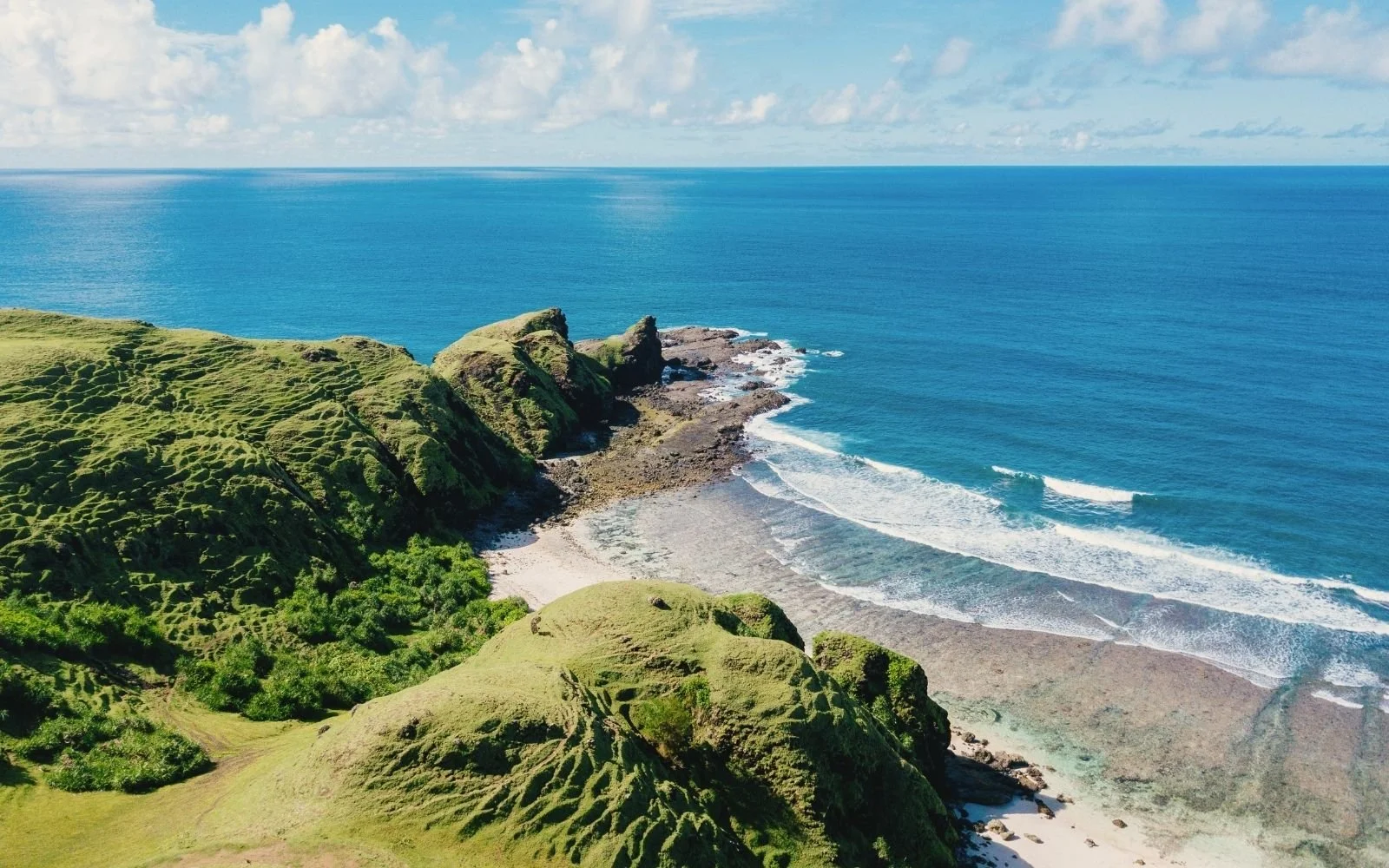 Aerial view of a lush green coastal landscape with rocky cliffs, a sandy beach, and the blue ocean extending to the horizon.
