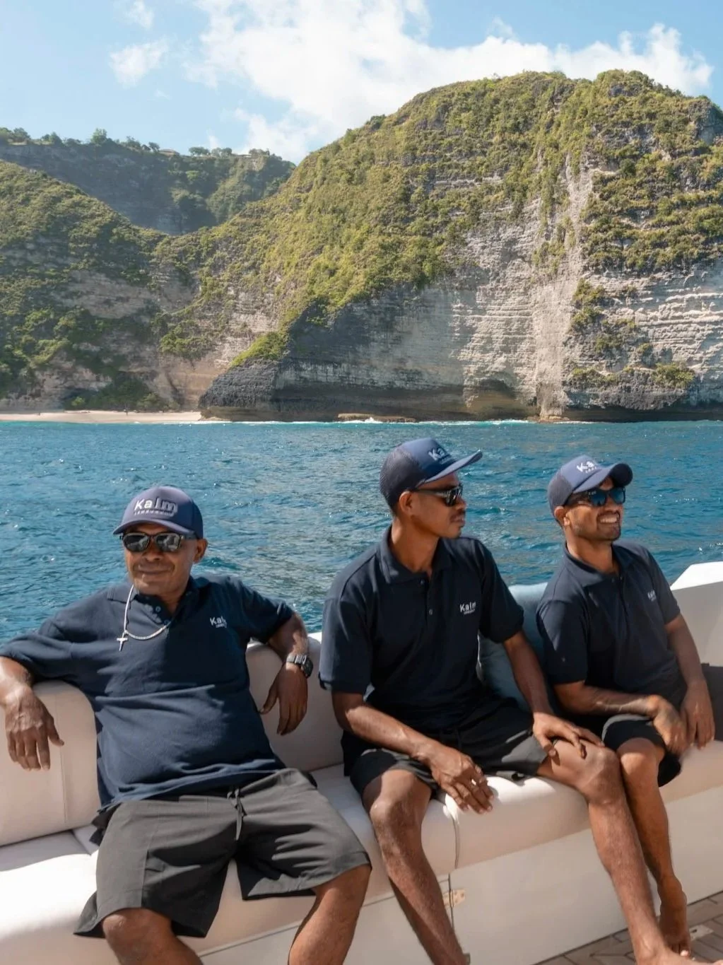 Three men sitting on a boat with a rocky green island in the background and blue ocean water.