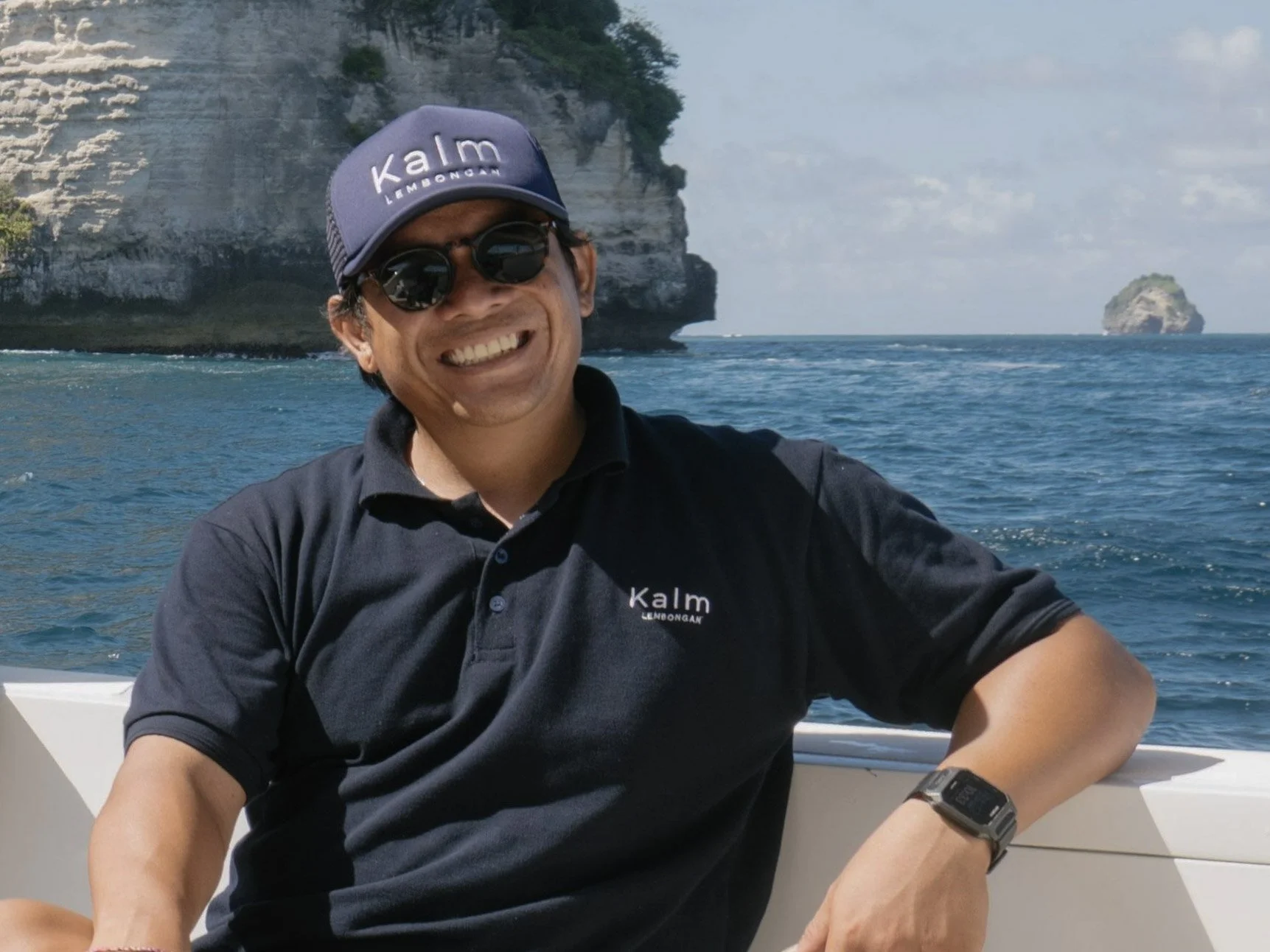 A smiling man wearing sunglasses and a navy blue cap with the words 'Kalm Lemonade' on it, posing on a boat with ocean water and rocky cliffs in the background.