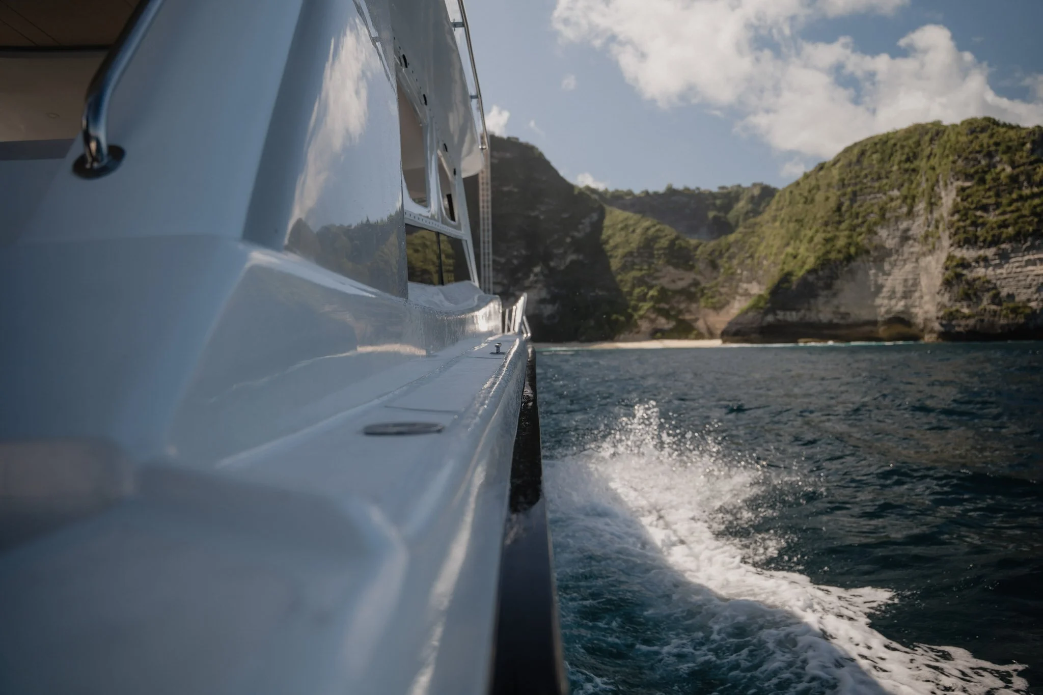 Photo taken from the side of a boat sailing in the water near green rocky cliffs under a partly cloudy sky.