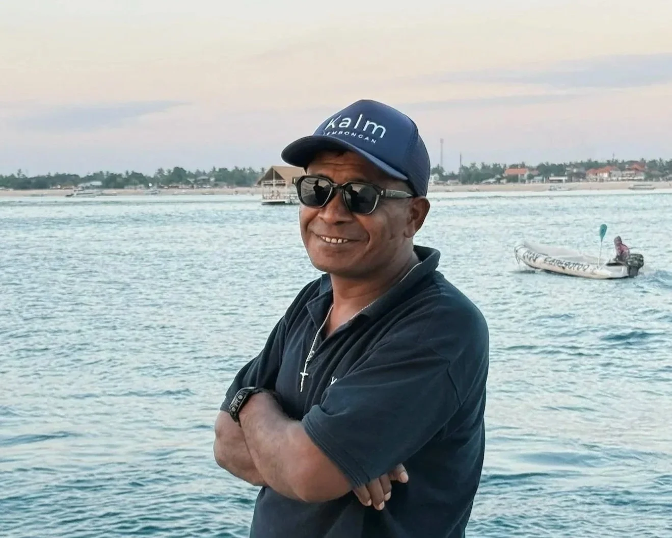 A man smiling with arms crossed, wearing sunglasses and a blue cap, standing by water with a kayak and a woman in the background.