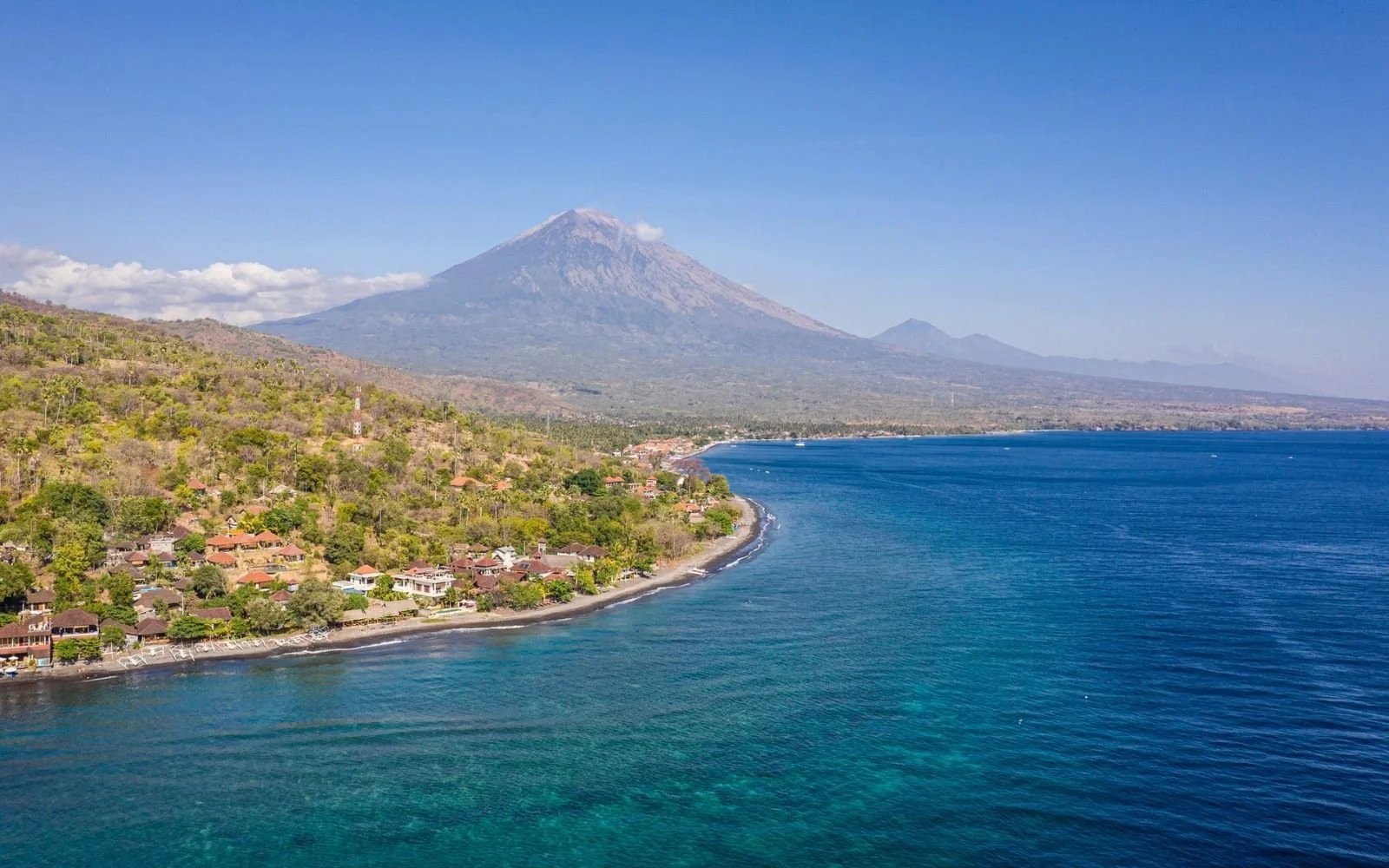 A tropical coastline with colorful houses and lush green hillside leading to an active volcano in the background, next to clear blue waters under a sunny sky.