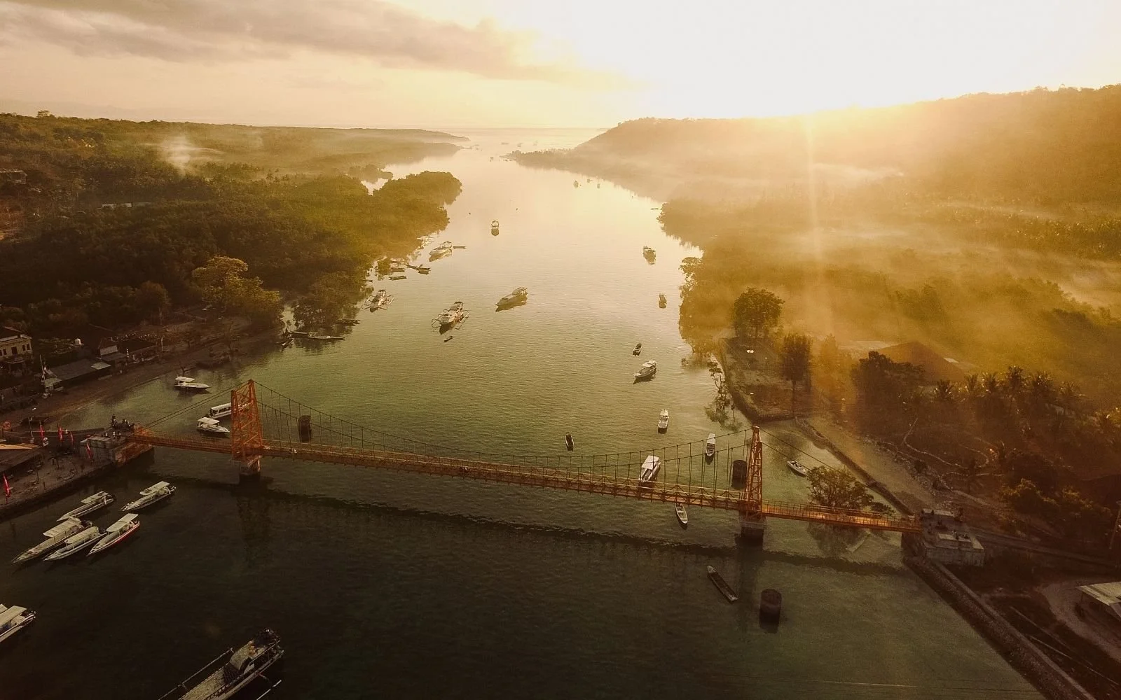 Aerial view of a river with boats, a bridge, and surrounding greenery during sunset.
